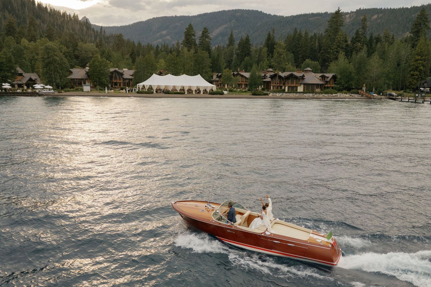 Vintage wooden boat cruising across lake with mountain landscape and white tent visible on shoreline.