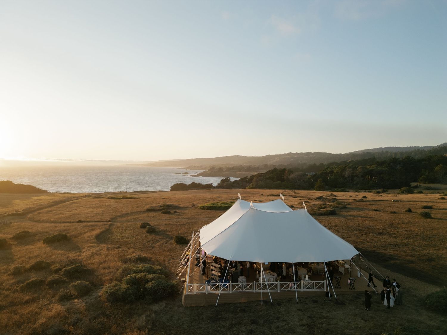 Large white wedding tent set up on coastal bluff at sunset with ocean views.