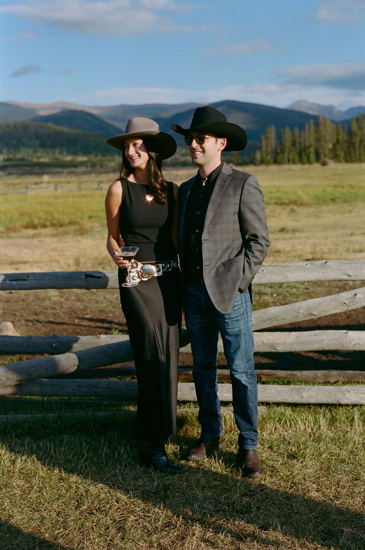 Rustic western-themed wedding portraits by a wooden fence in a scenic mountain meadow.