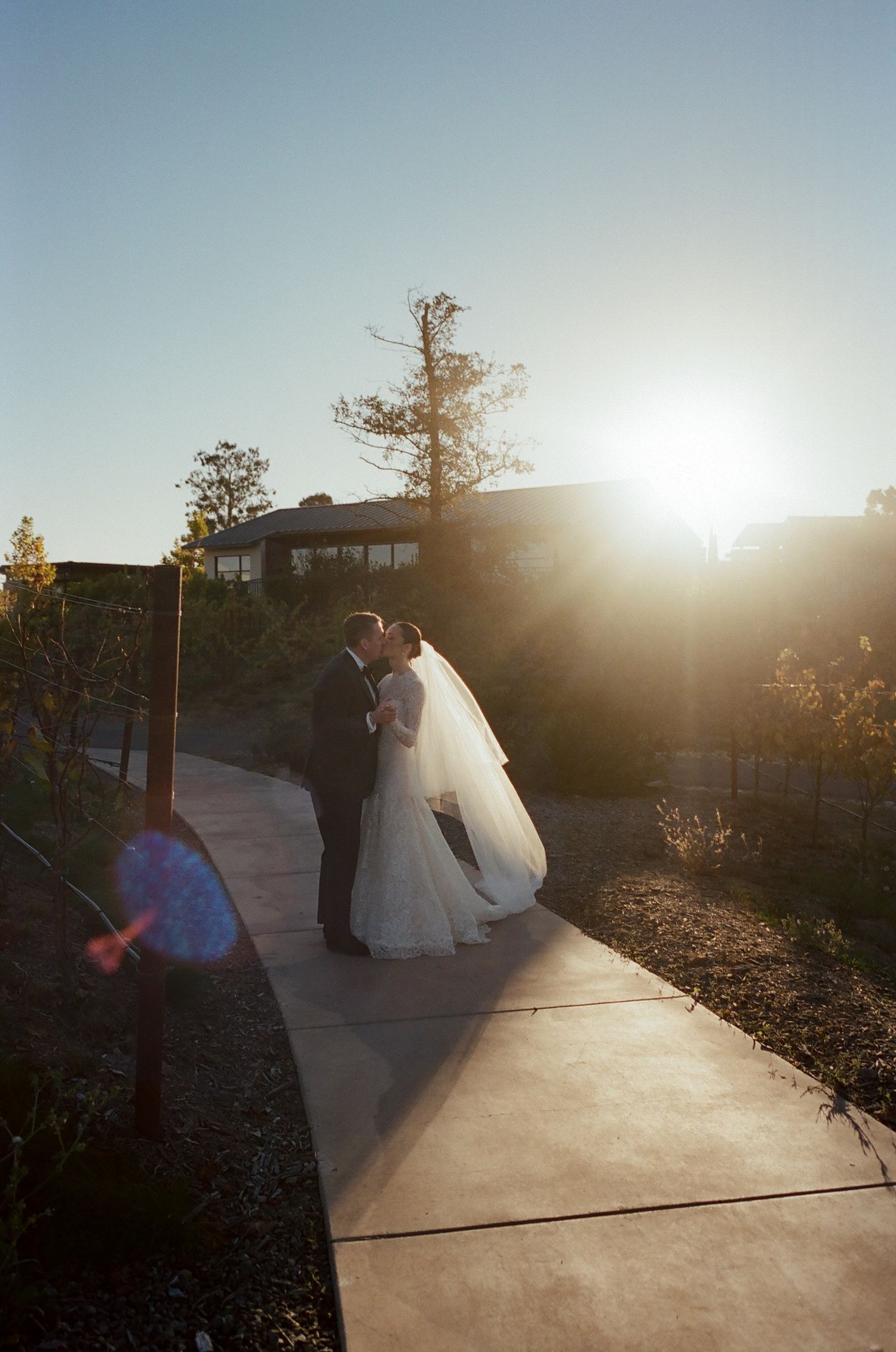 Wedding couple silhouetted against sunset on concrete path with lens flare.