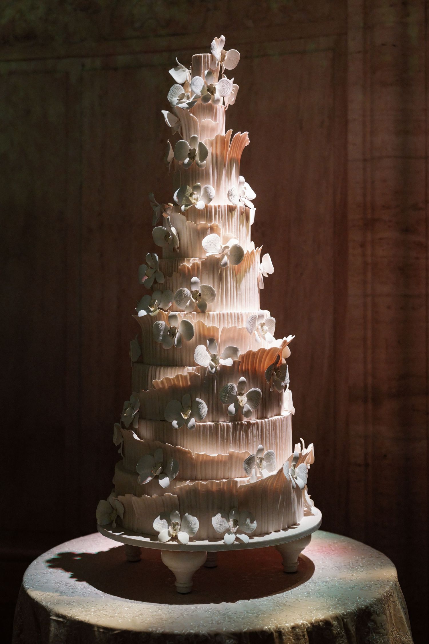The image shows a large, multi-tiered wedding cake decorated with flowers and lights, sitting on a table.