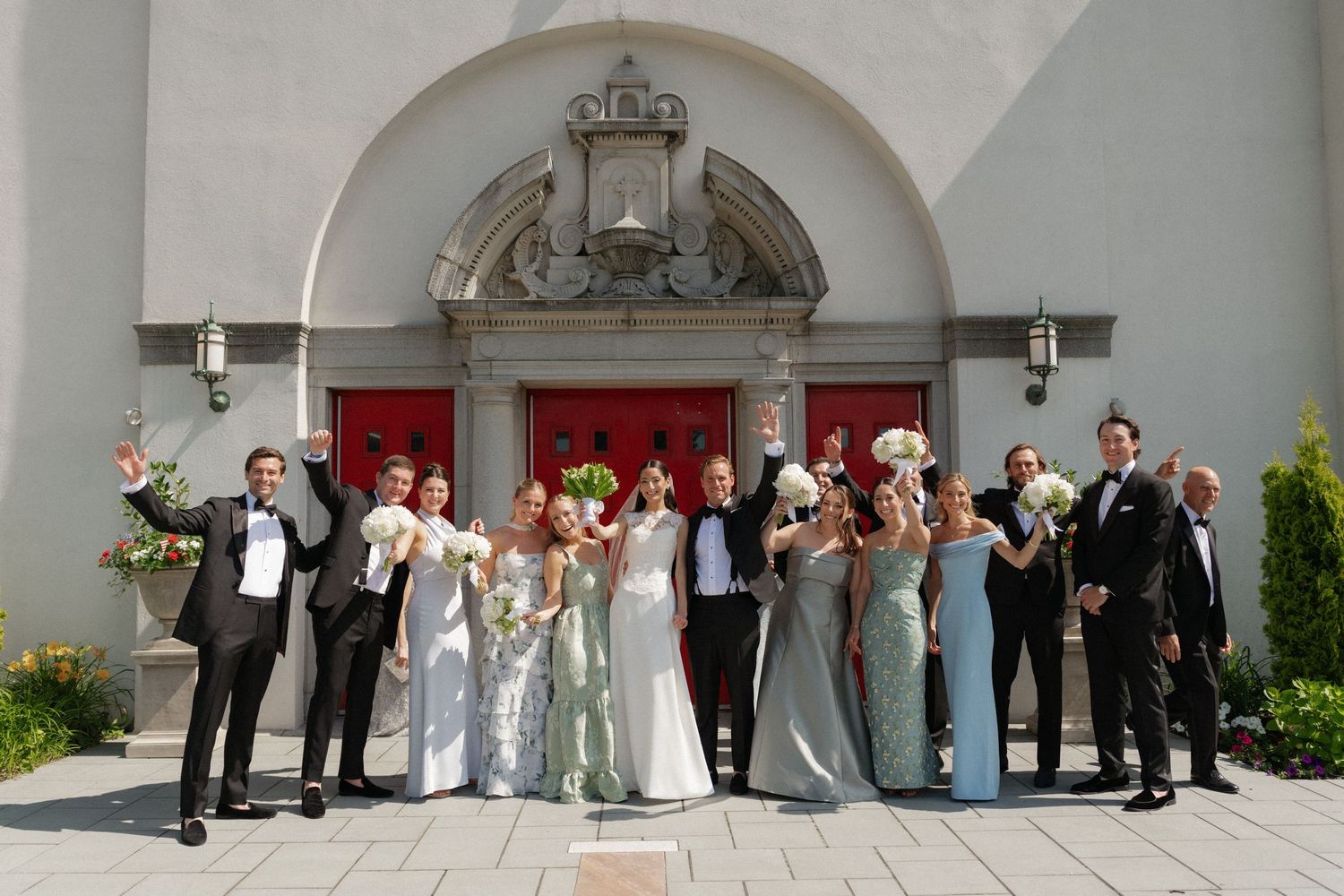Wedding party celebrating outside a church with red doors, bridesmaids in sage gowns and groomsmen in tuxedos.