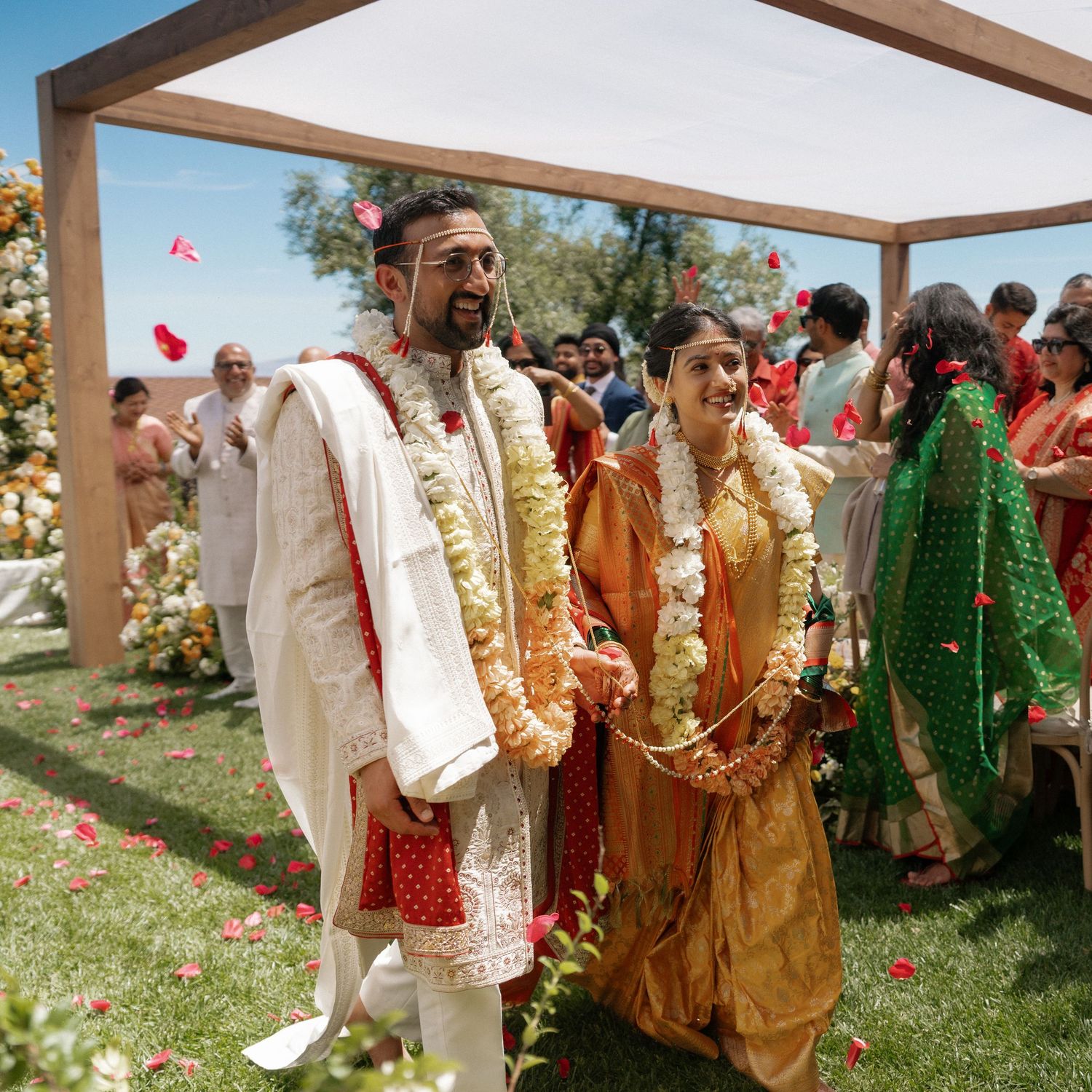 Indian bride and groom in traditional attire walk under floral mandap during outdoor Hindu wedding ceremony.