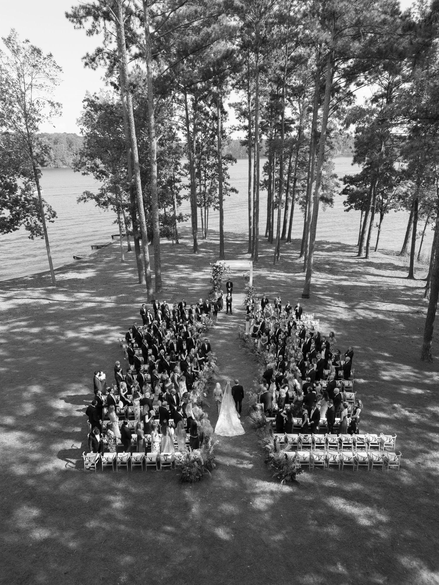 Aerial view of wedding ceremony with guests arranged in a heart shape among pine trees.