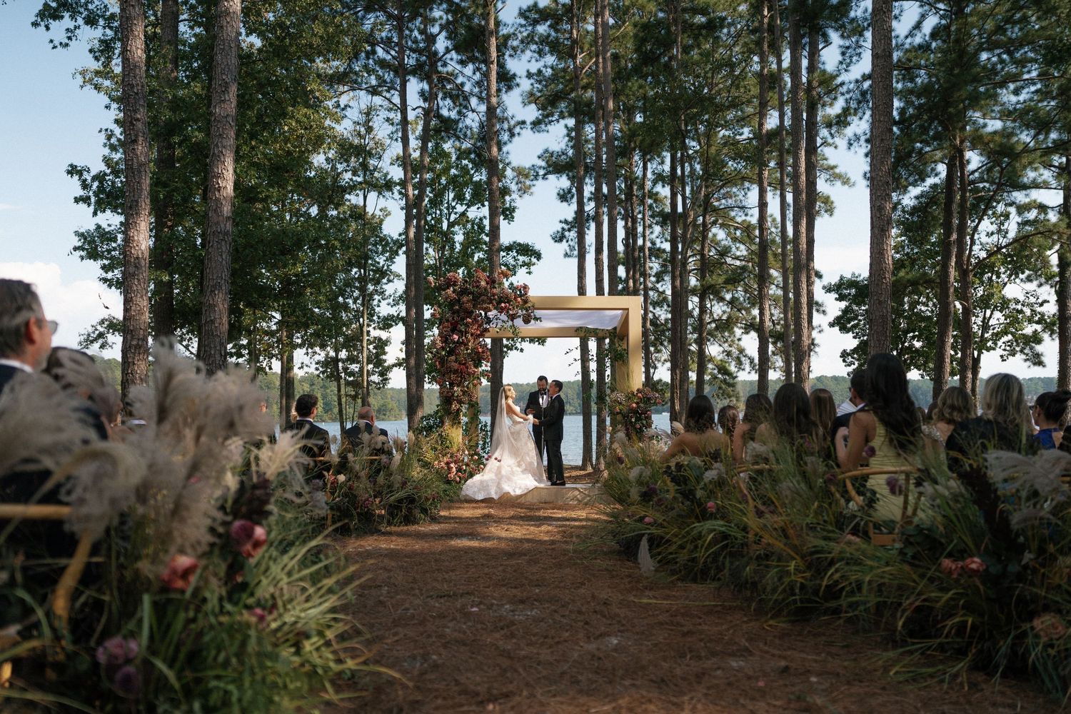 Wedding guests seated in white chairs between tall pine trees overlooking a serene lake.
