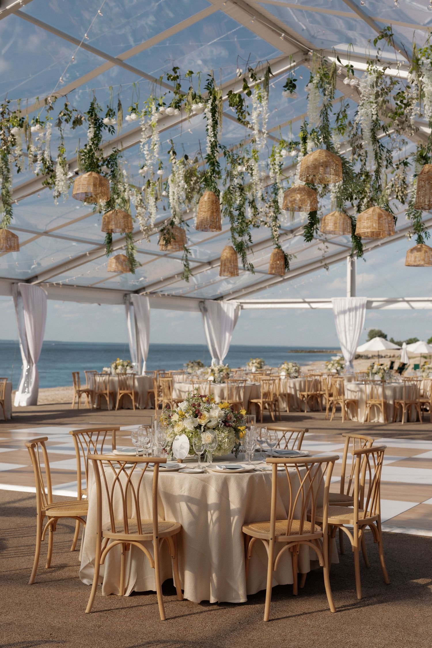 Elegant beachside wedding reception under clear tent with hanging greenery, wicker lanterns, and gold chiavari chairs.