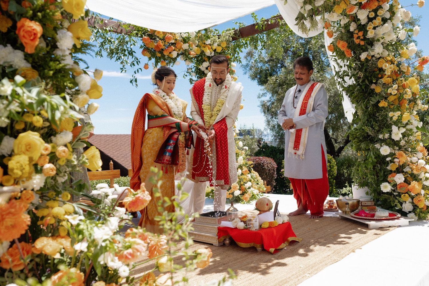 Couple exchanges vows at vibrant Indian wedding ceremony under floral arch with yellow and orange blooms.