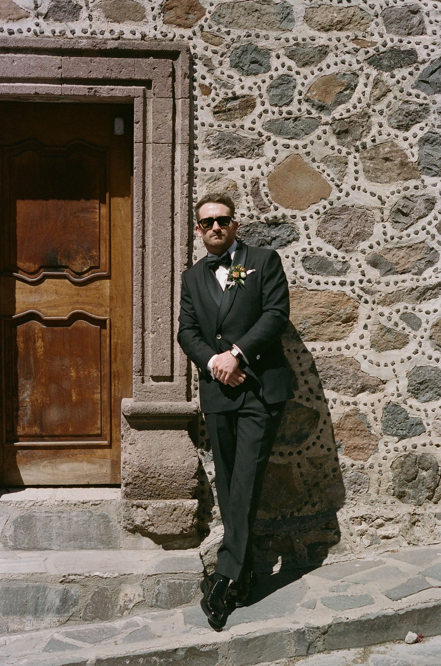 Formal attire and sunglasses against rustic stone wall with wooden door.