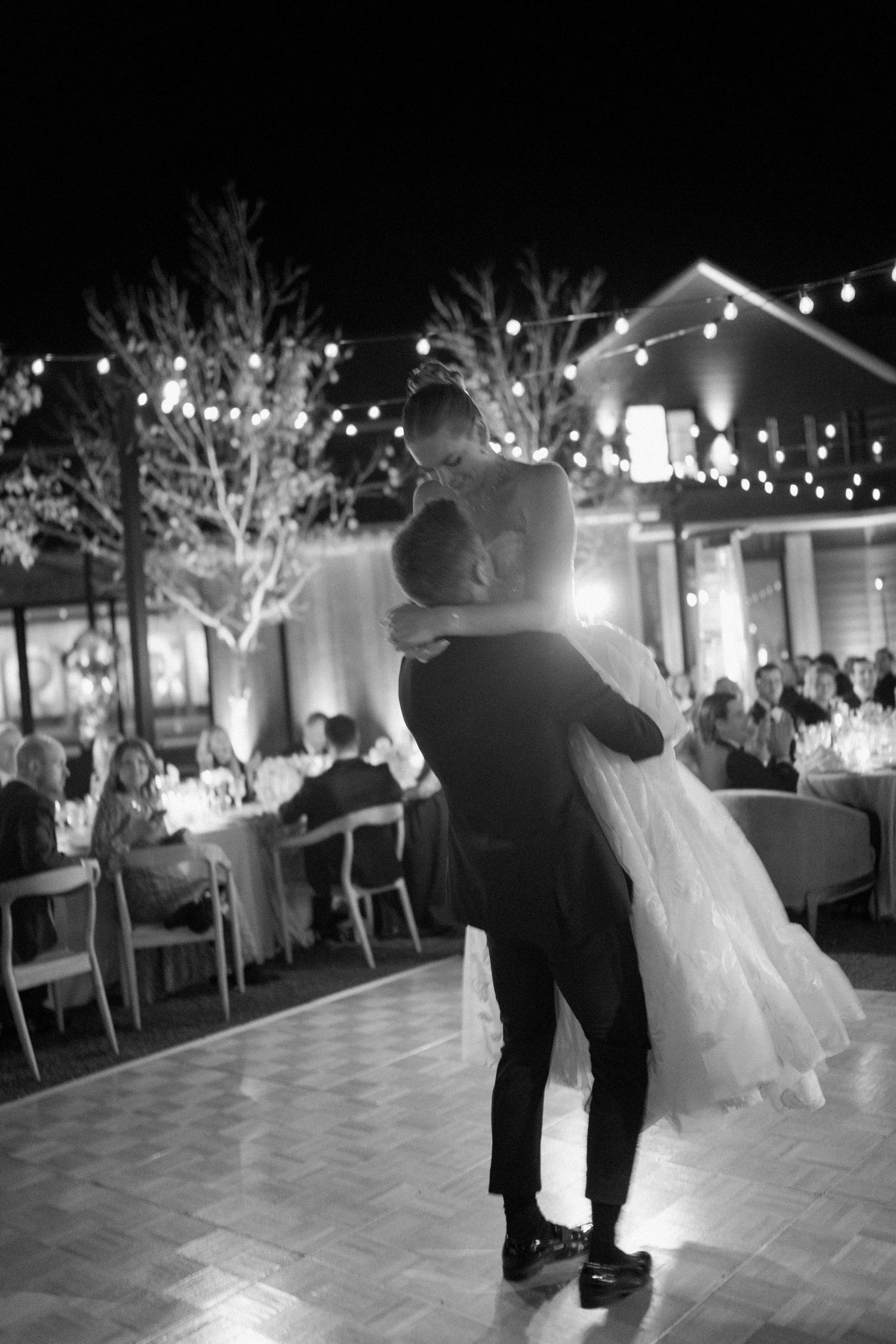 A couple dancing at an outdoor wedding reception under twinkling lights.