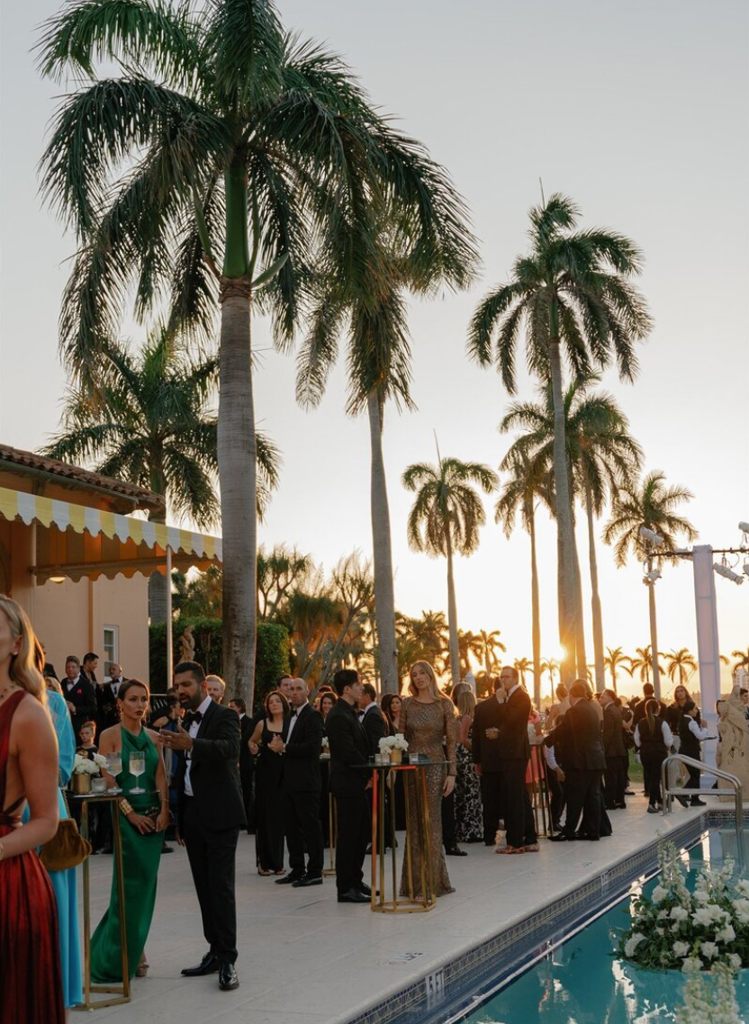 Wedding guests mingle at an outdoor reception by a pool with palm trees at sunset.