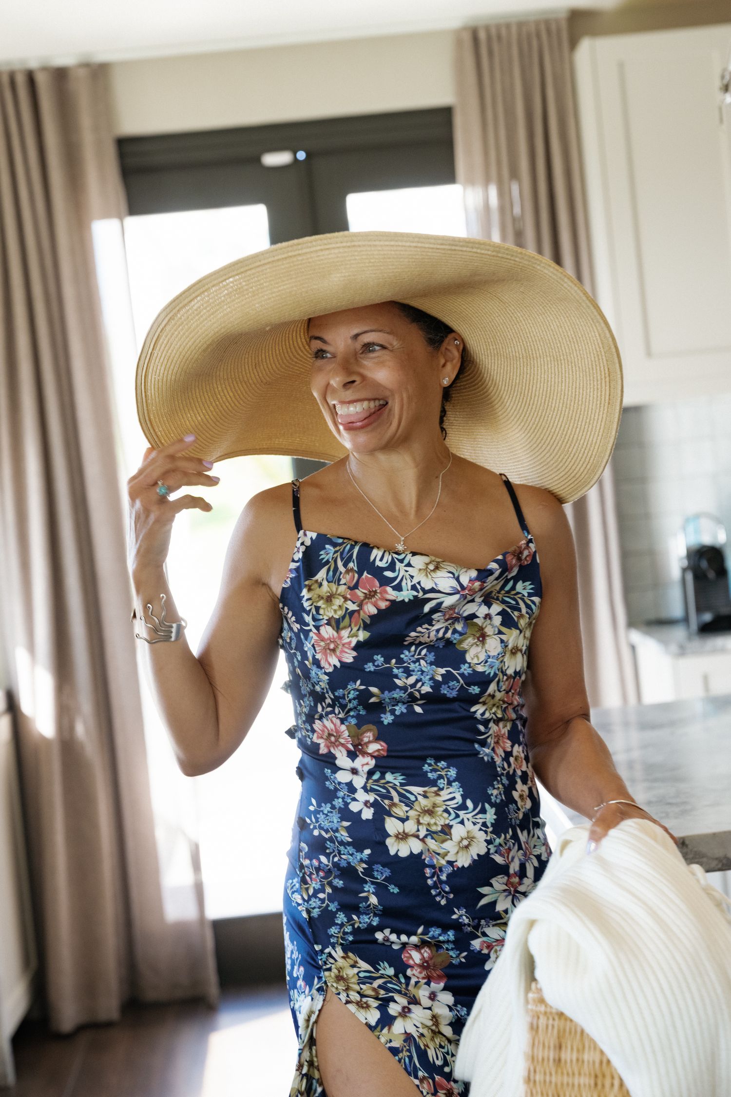A smiling woman wearing a floral dress and a large sun hat stands in a room with curtains.