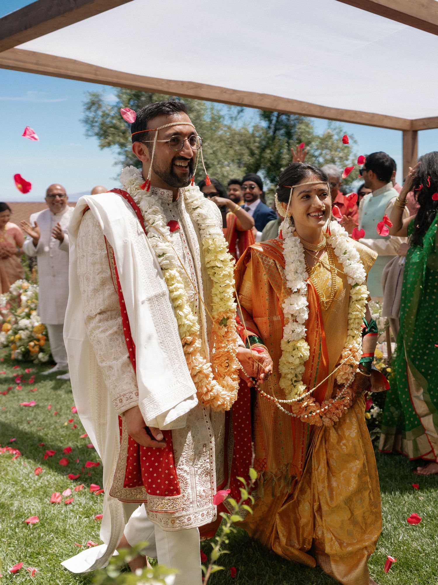 Indian wedding couple in traditional attire walking under floral shower, bride in gold saree and groom in white sherwani.