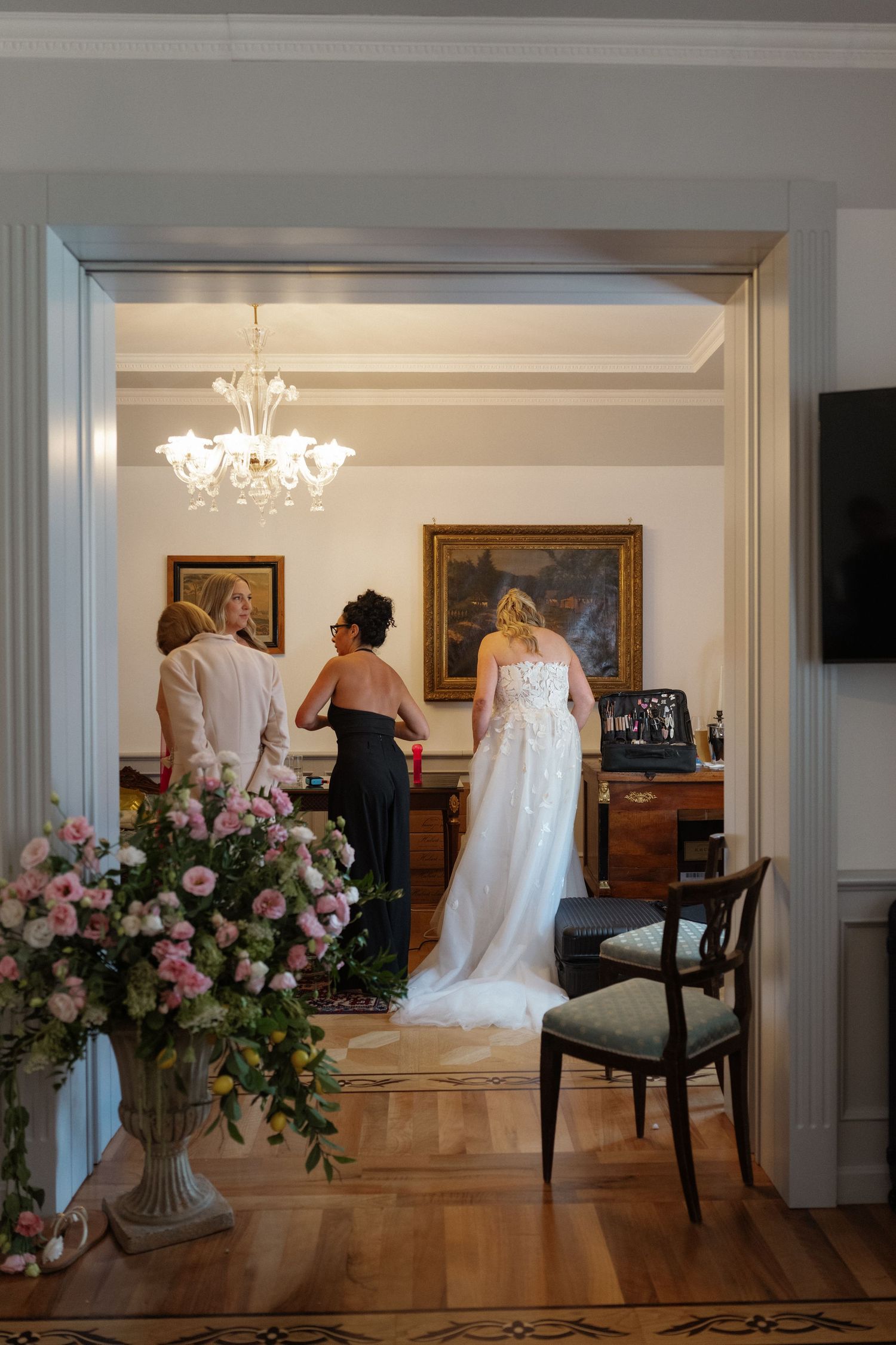 Bride in white wedding gown stands with guests in elegant room with chandelier and pink floral arrangement.