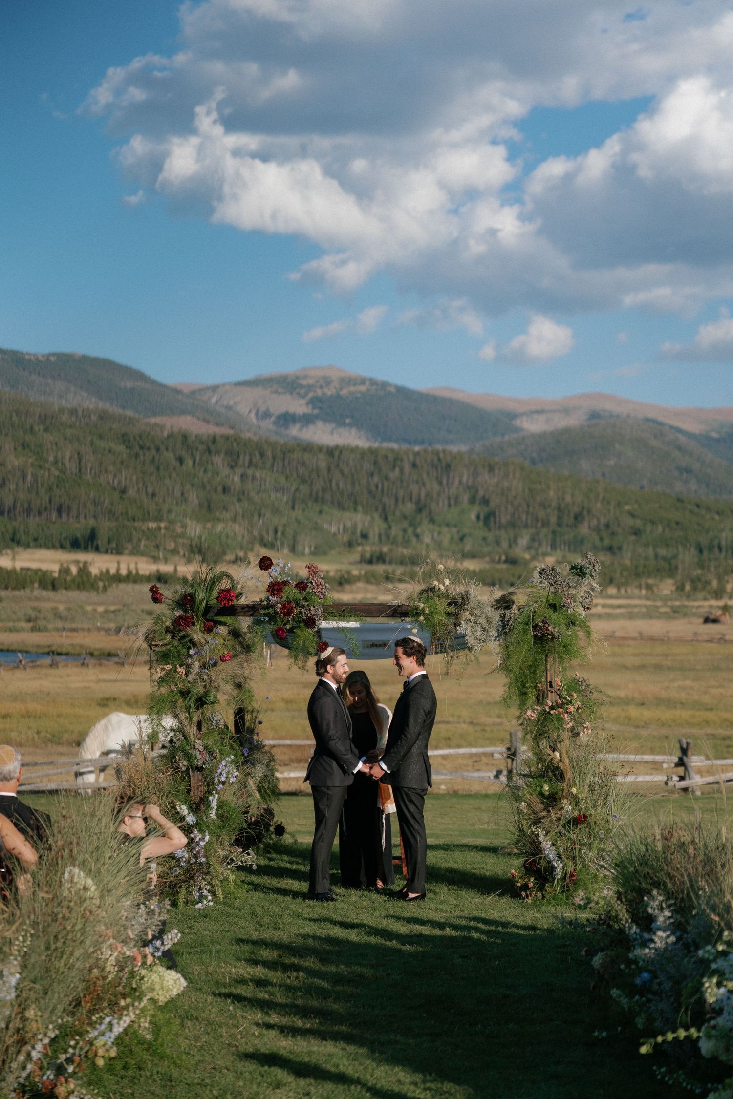 Outdoor wedding ceremony setup with mountain backdrop and floral arch arrangements in a scenic meadow.