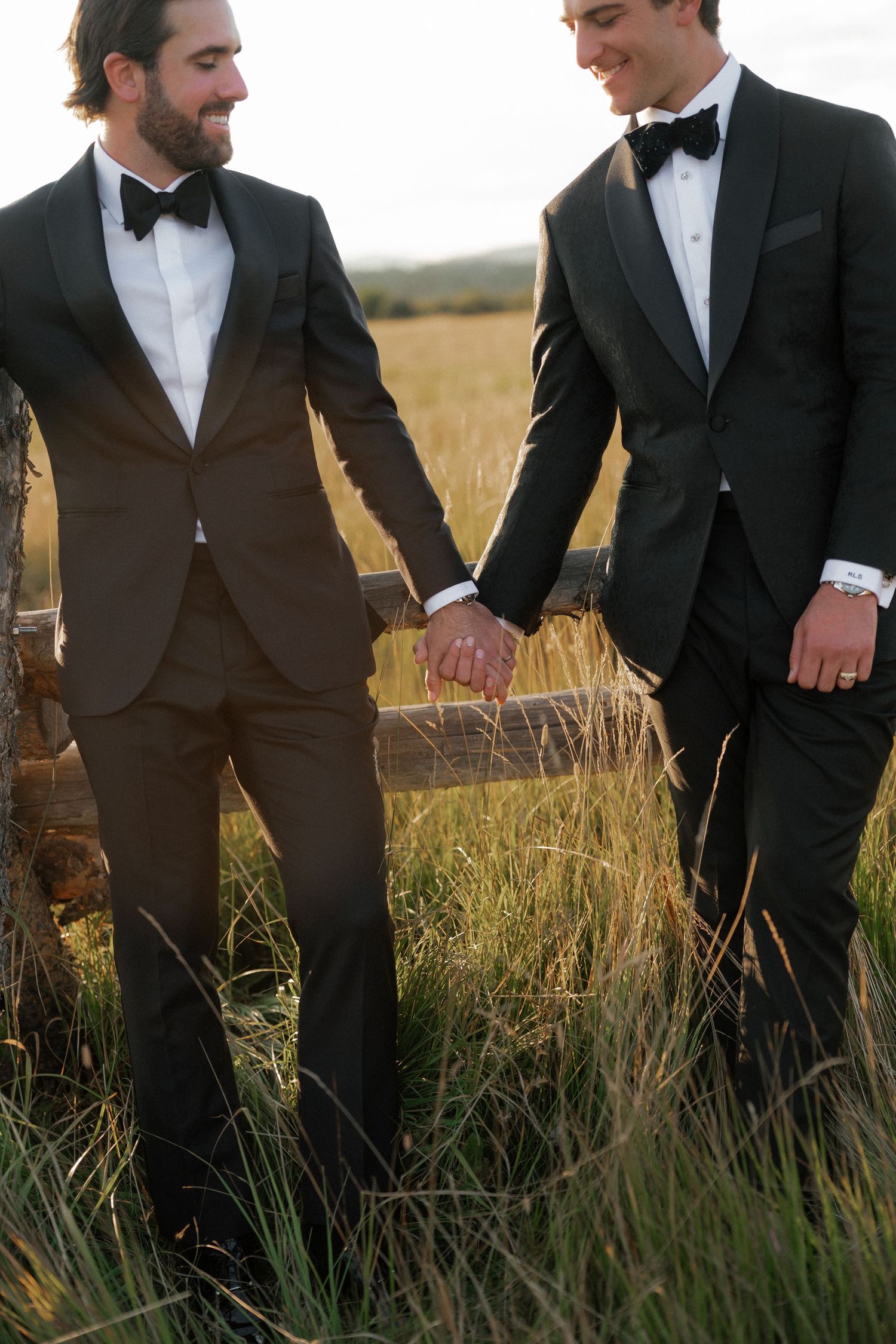 Groomsmen in black tuxedos stand together in a golden field at sunset.