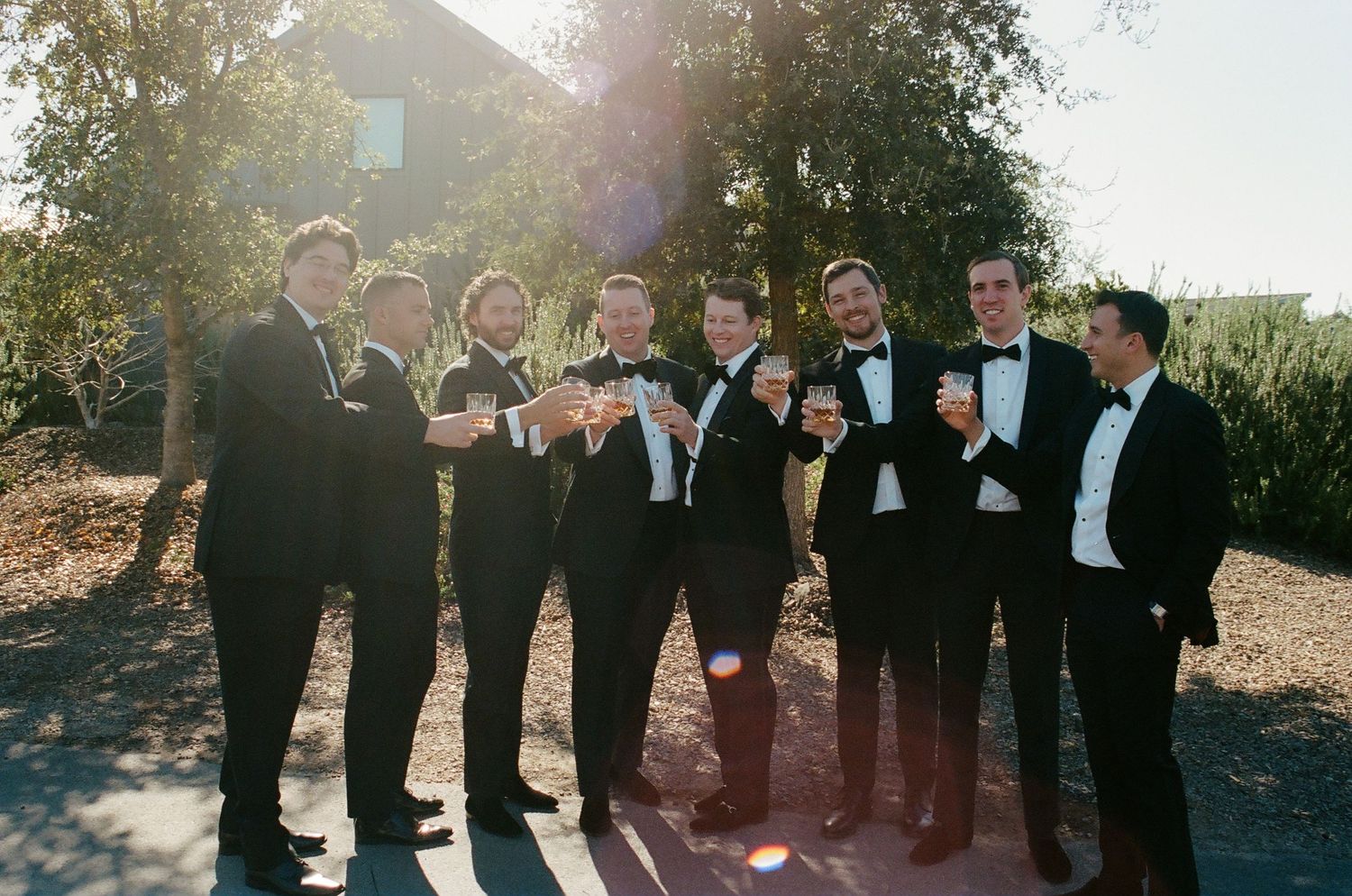 Groomsmen in black tuxedos pose together outdoors for formal wedding photos.