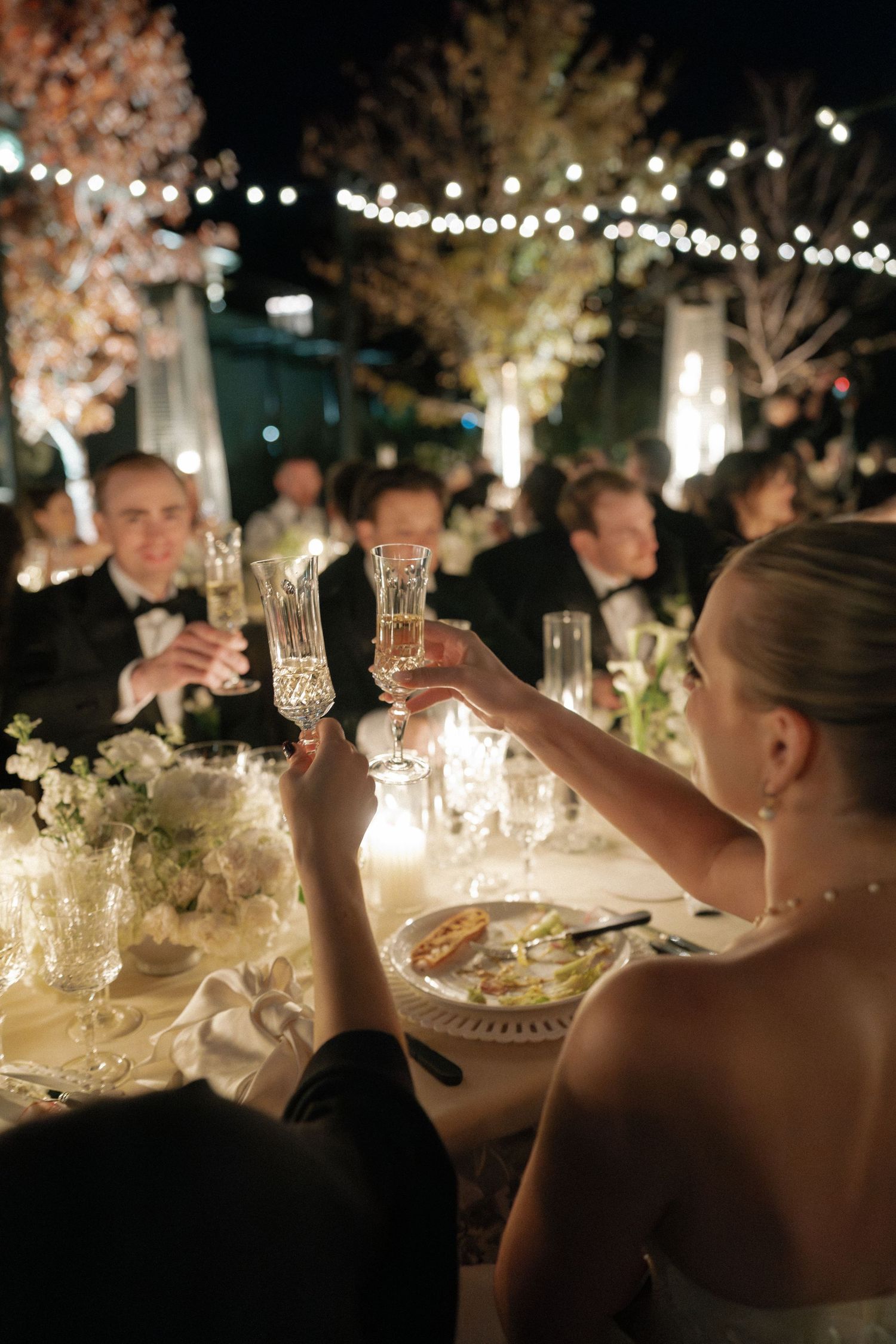 Wedding guests raise glasses for a toast at an elegant outdoor reception with string lights.