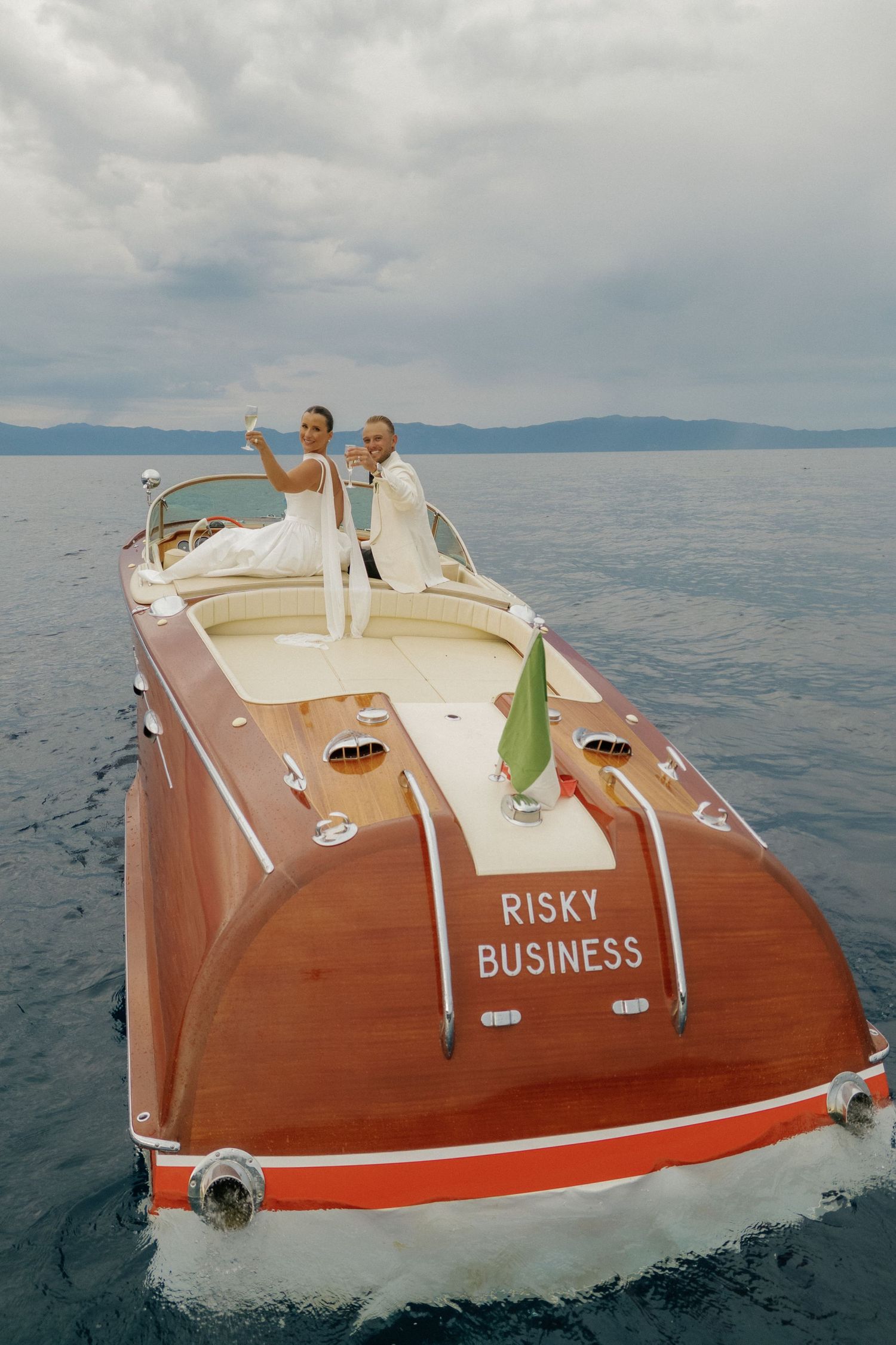 A couple stands on a vintage wooden boat named 'Risky Business' on a serene lake.