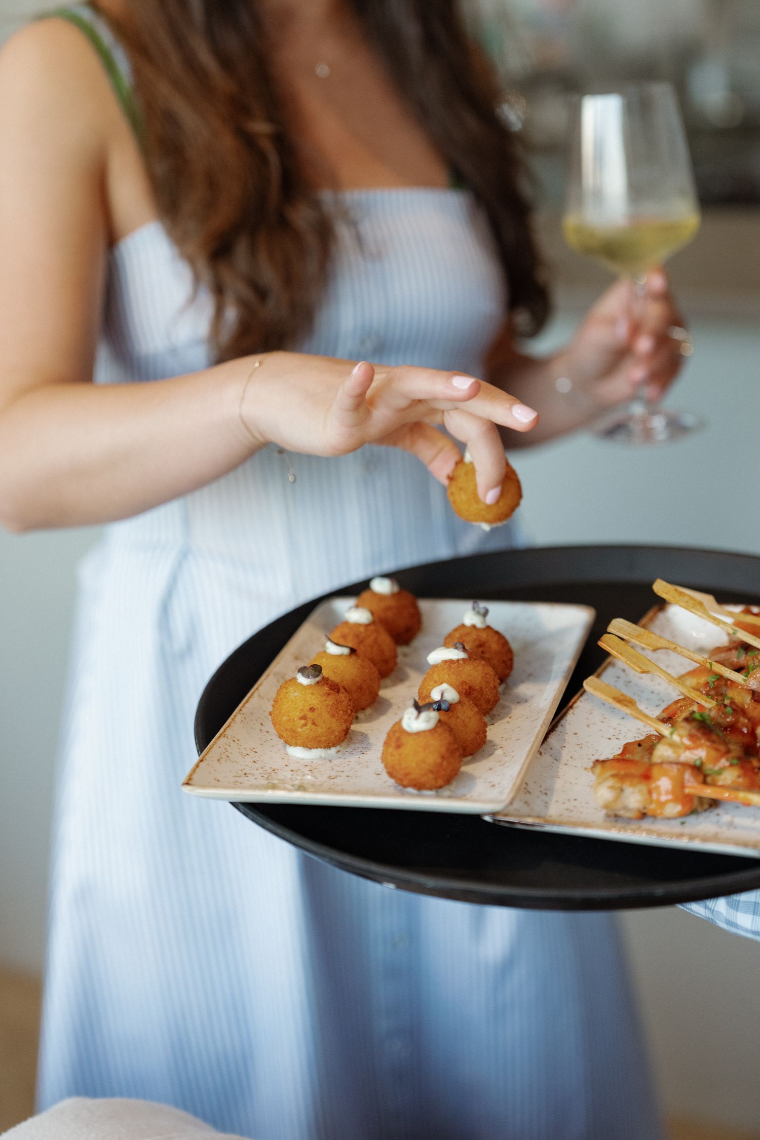A woman in a light blue dress holds a plate of fried appetizers and a glass of white wine.