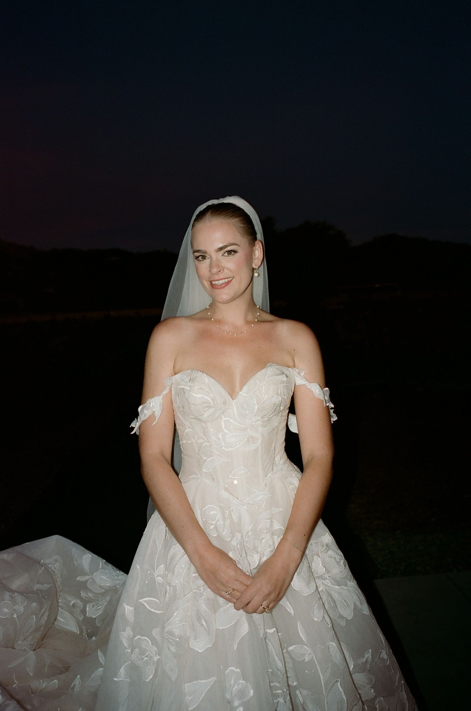 A bride wearing a strapless lace ballgown with off-shoulder details poses at dusk.