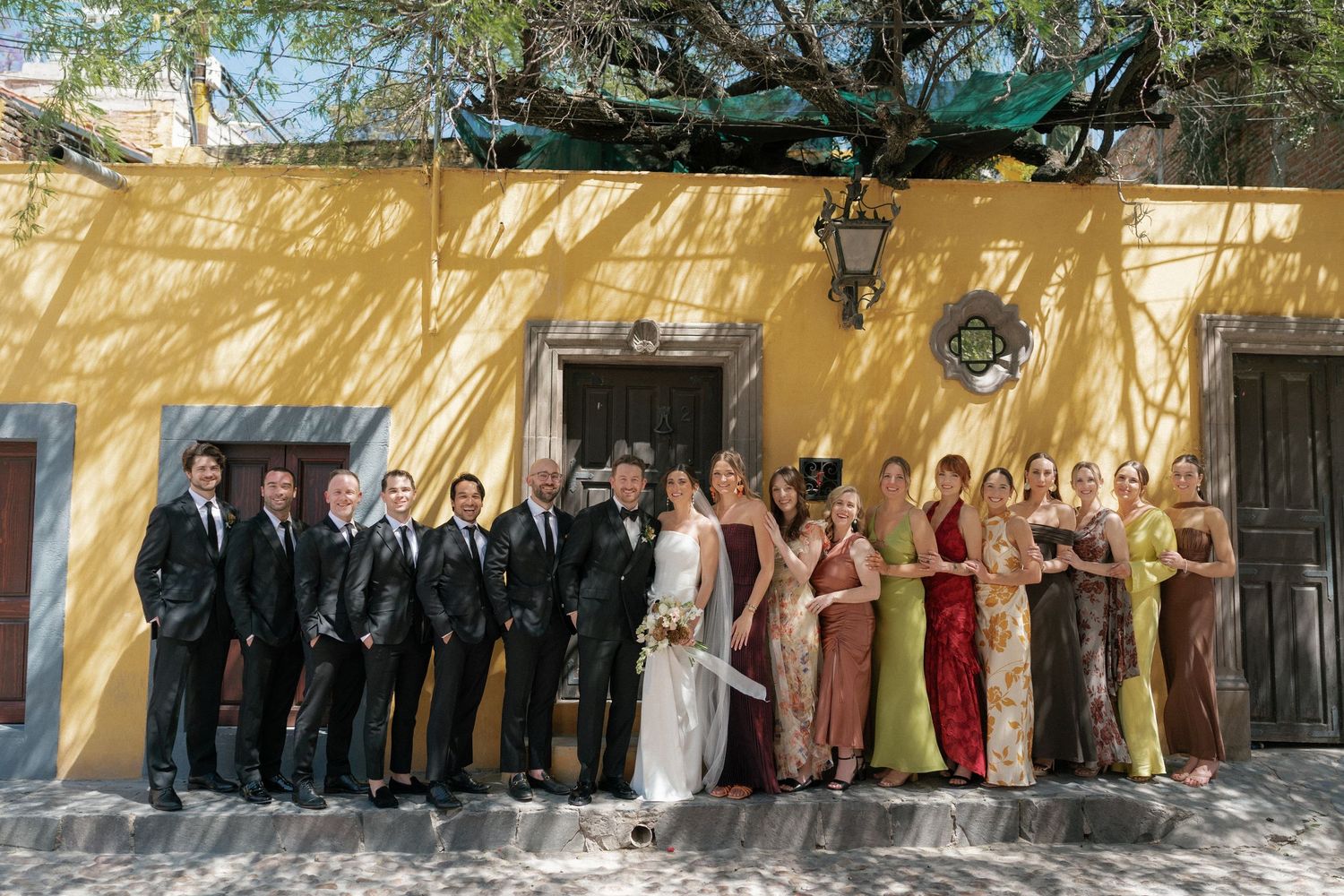 Wedding party lined up against a yellow wall for formal group photos.