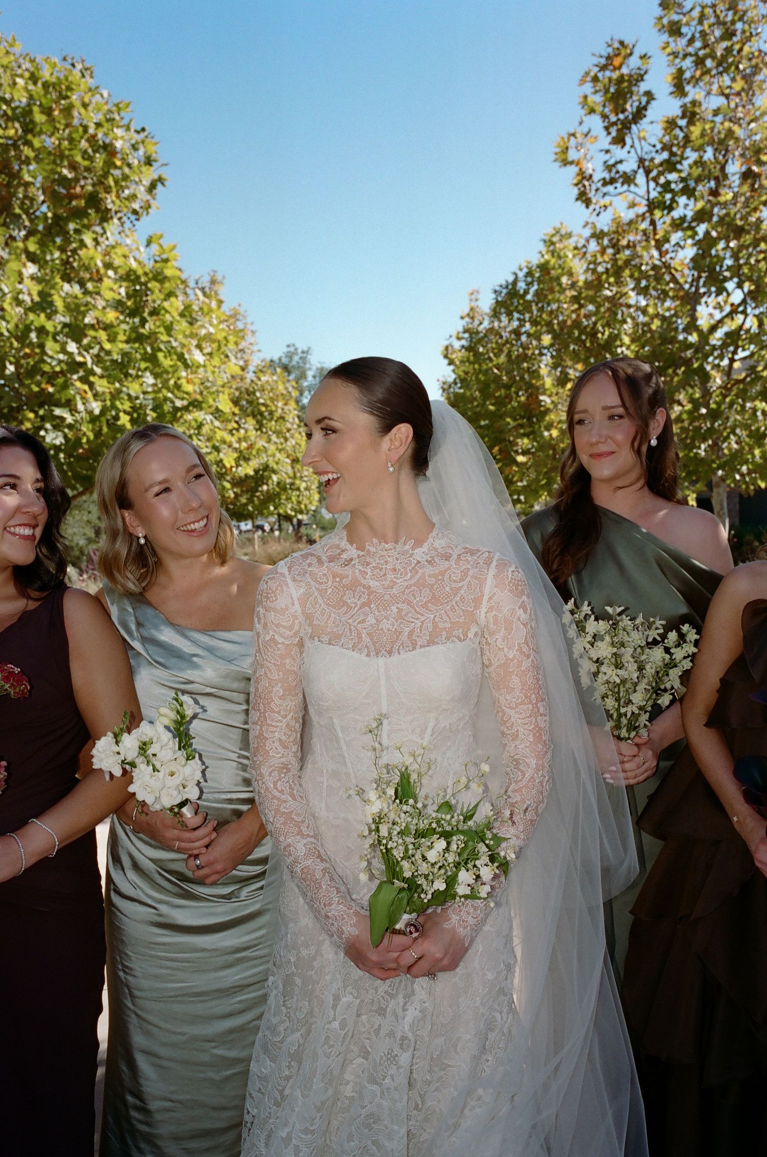Wedding party members in sage green and dark dresses gather outdoors under sunny skies.