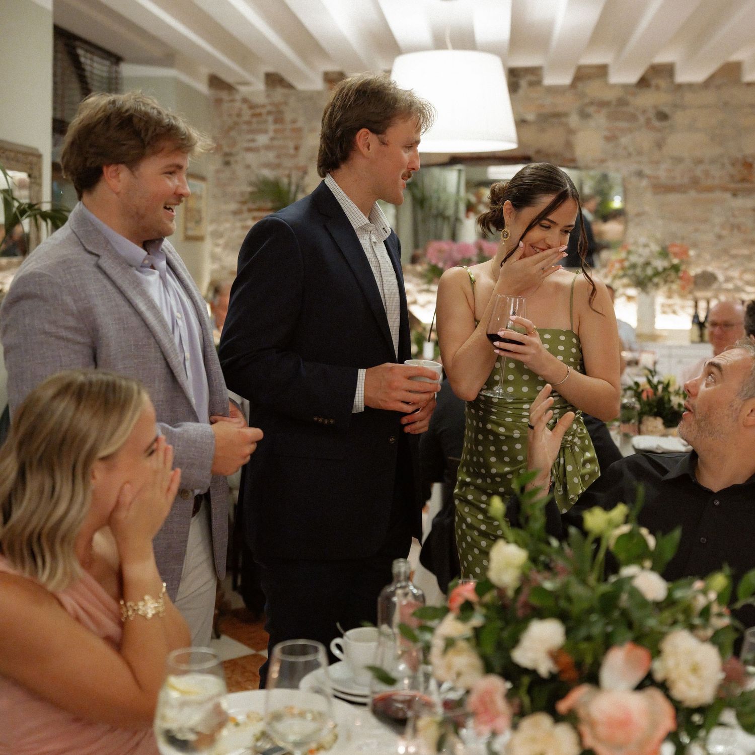 Guests laughing and socializing at an elegant wedding reception with floral centerpieces and rustic stone walls.