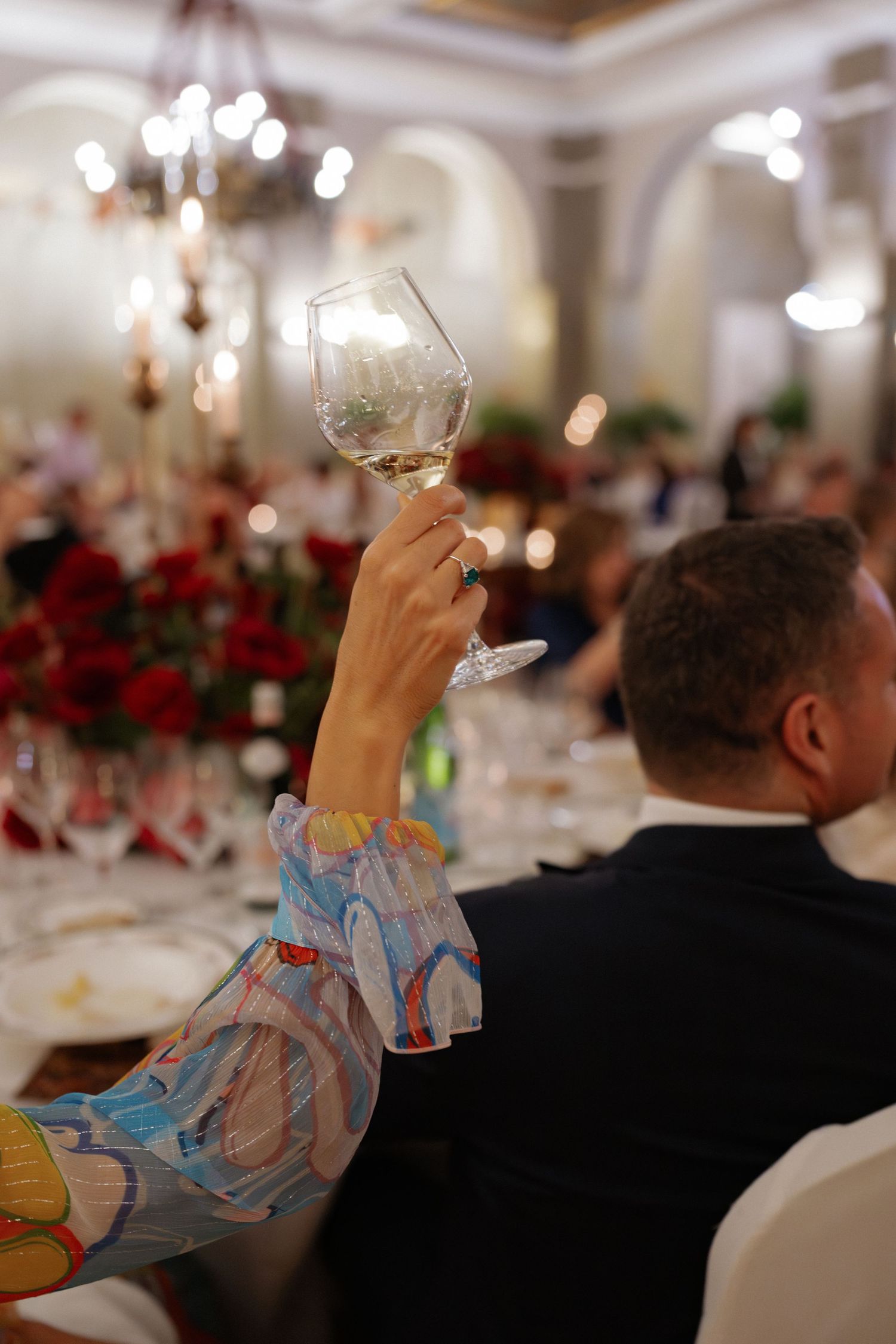 Woman in colorful floral dress raises wine glass in toast at formal gala dinner event.