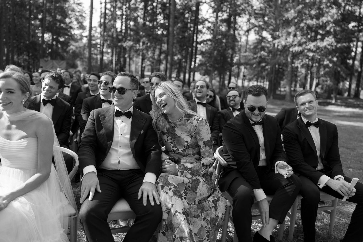 Wedding ceremony in progress with guests seated in wooden chairs under tall pine trees.