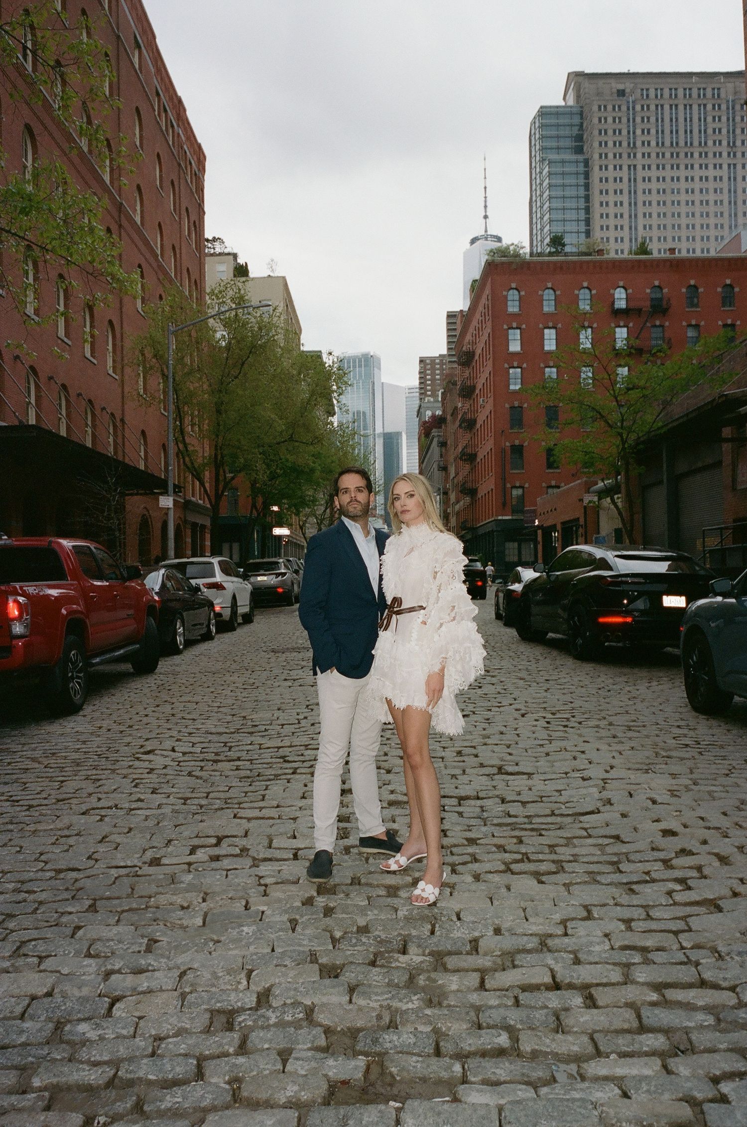 A couple poses on a cobblestone street lined with brick buildings and parked cars in an urban setting.