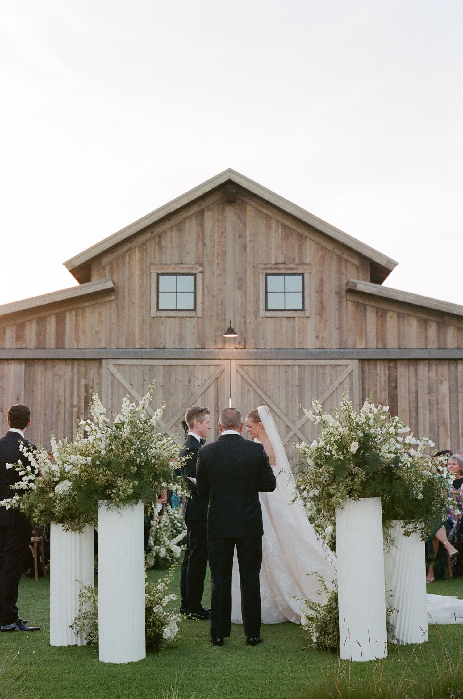 Wedding ceremony setup with white pillars and floral arrangements in front of a wooden barn.