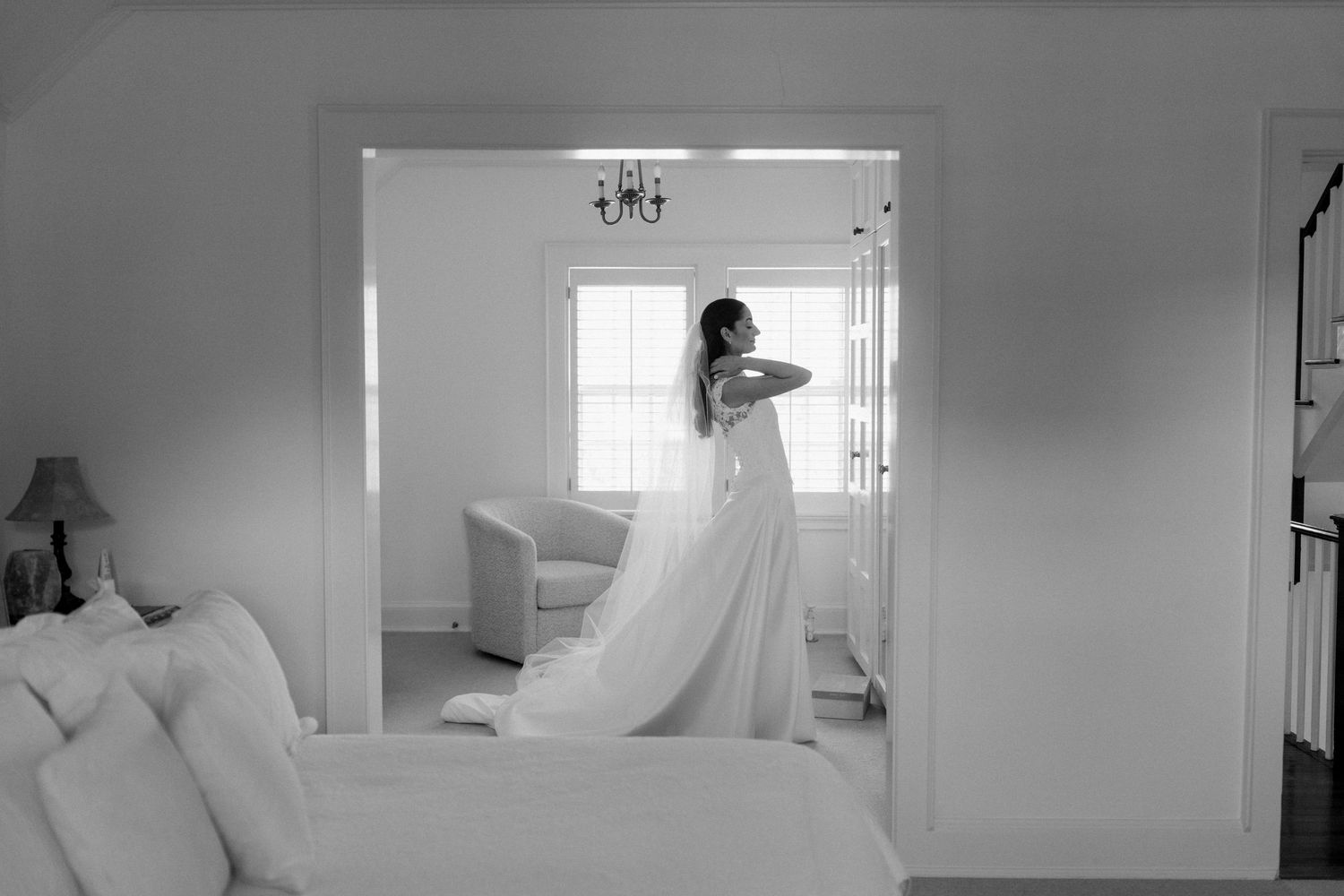 Black and white photo of a bride in a flowing wedding gown standing by a sunlit window in an elegant room.