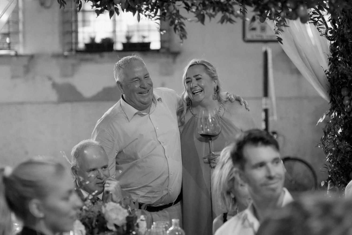 Happy couple smiling at an elegant indoor wedding reception with guests and floral arrangements.