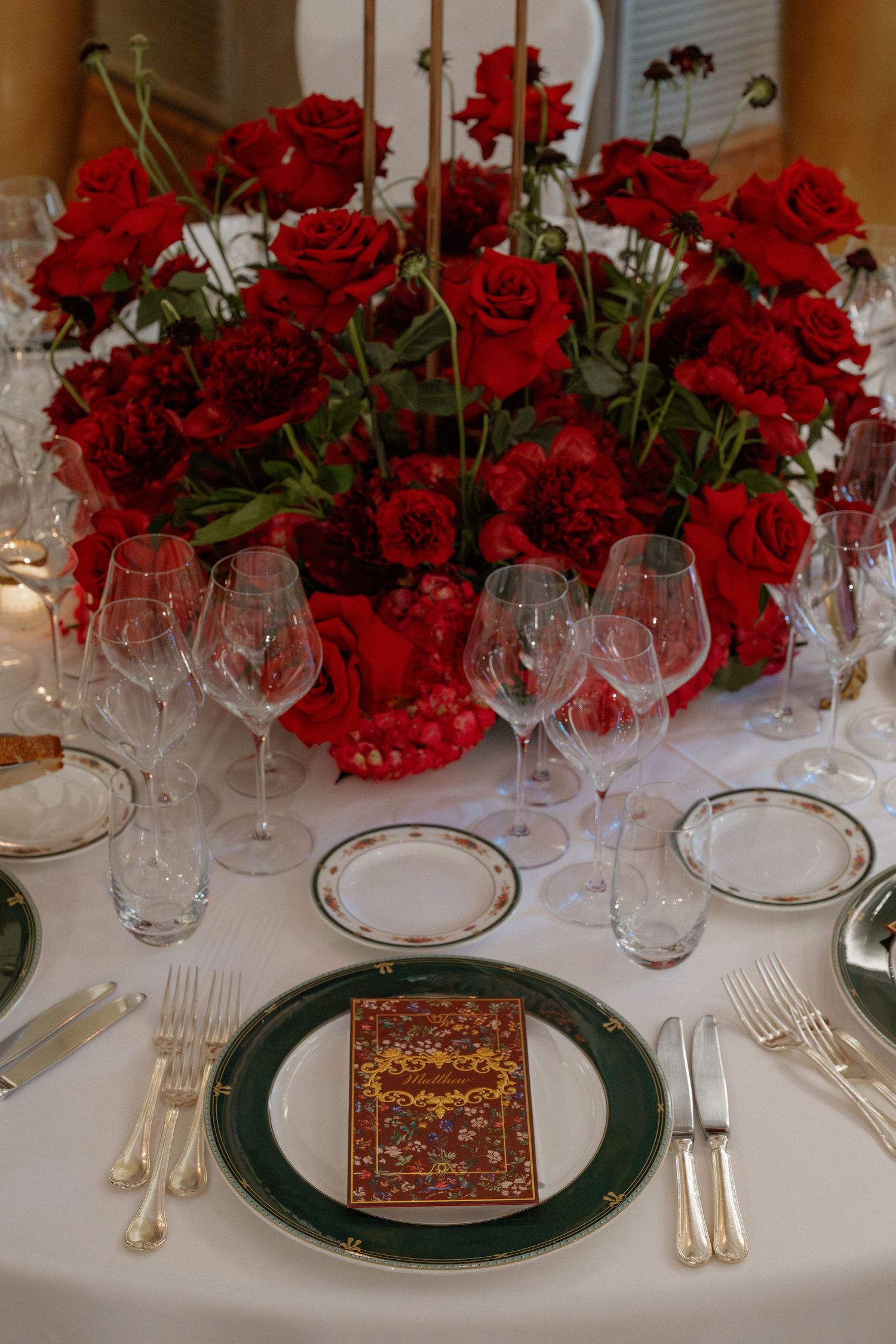 Elegant wedding table setting with red roses centerpiece, crystal glasses, and green gold-rimmed plates.
