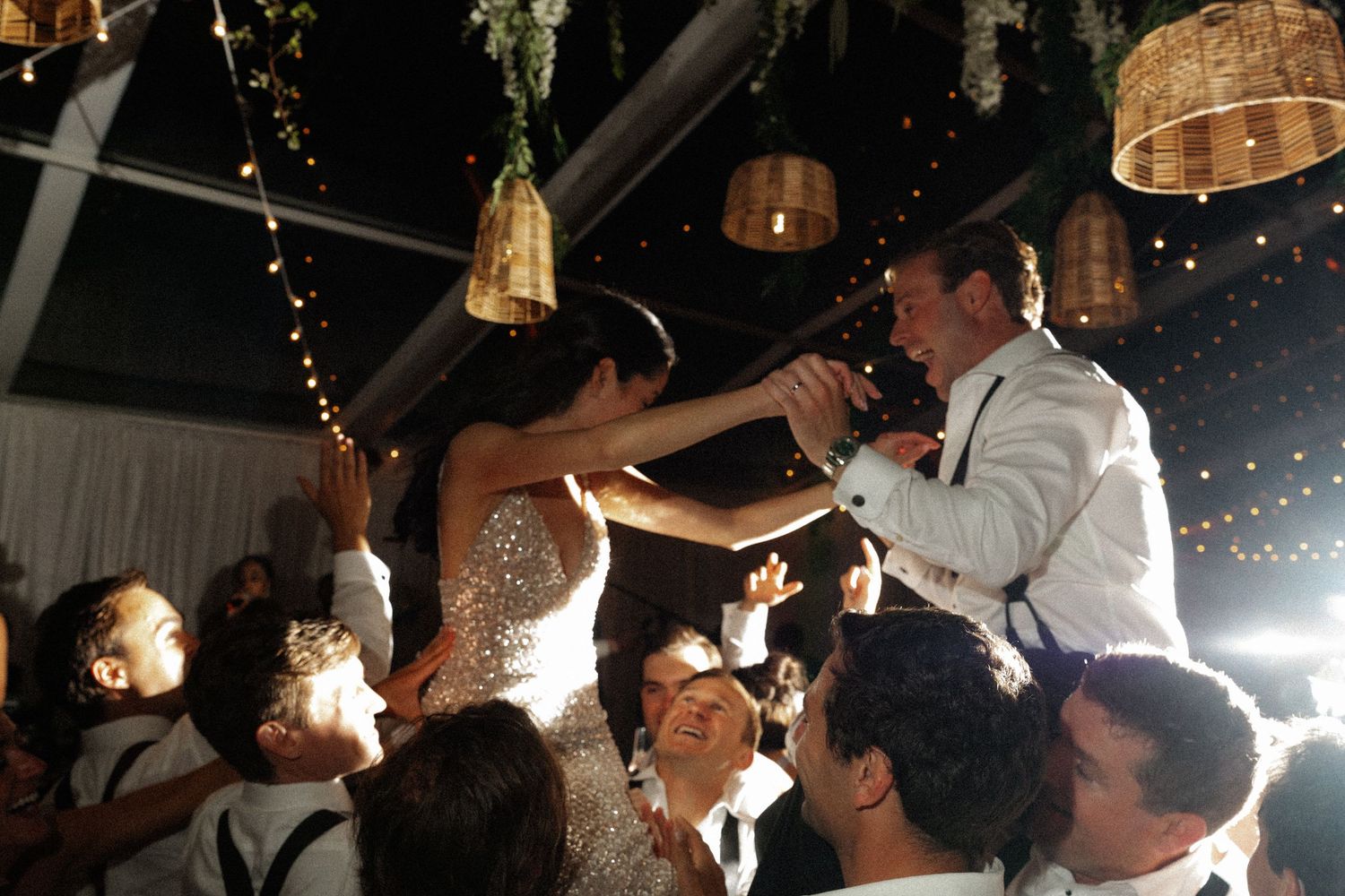 Bride and groom lifted by guests during lively wedding reception dance under wicker pendant lights.