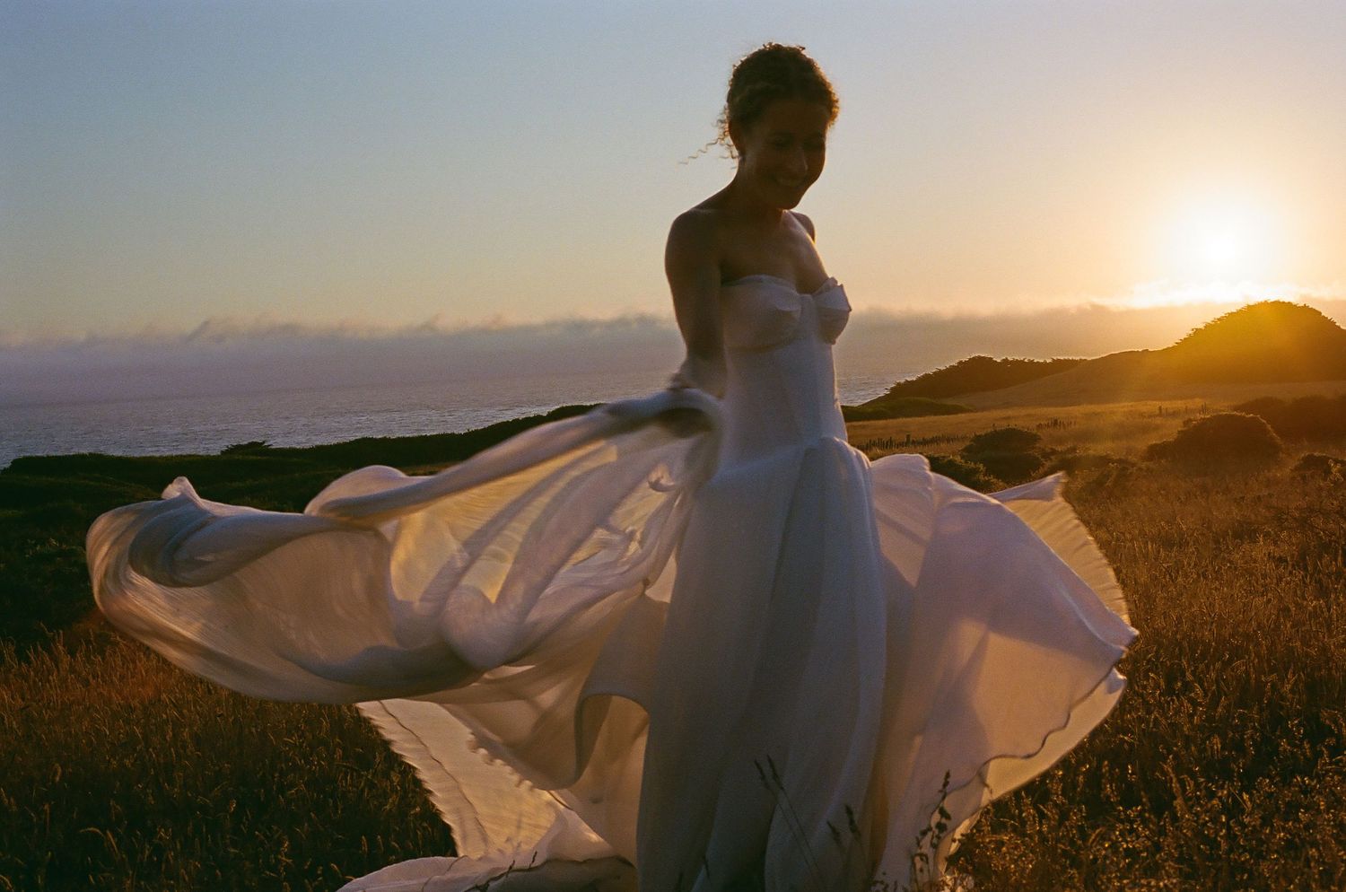 White wedding dress flowing dramatically against golden sunset in coastal field setting.