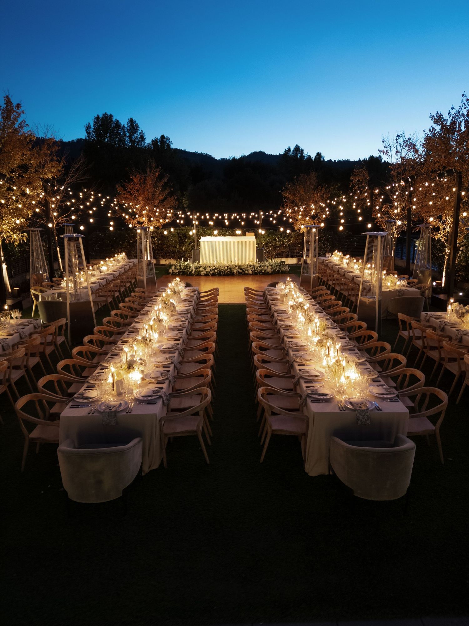 Long banquet tables set for an outdoor evening wedding reception with twinkling lights.
