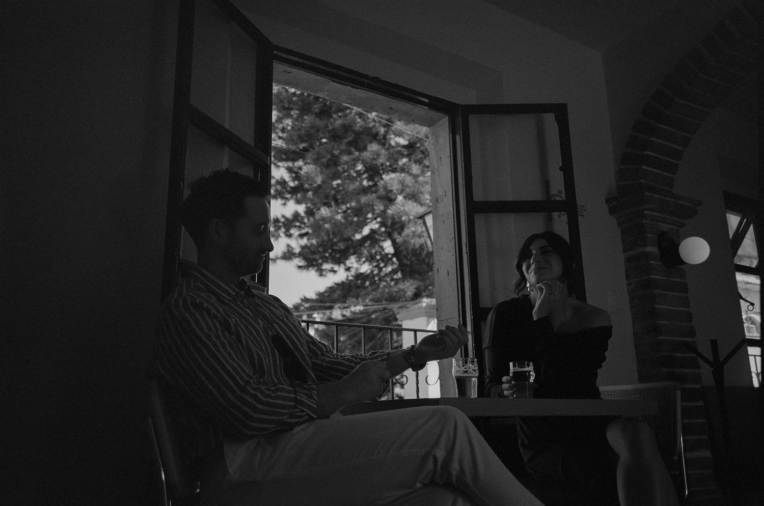 Black and white photograph of a silhouetted figure sitting by an open window with trees visible outside.