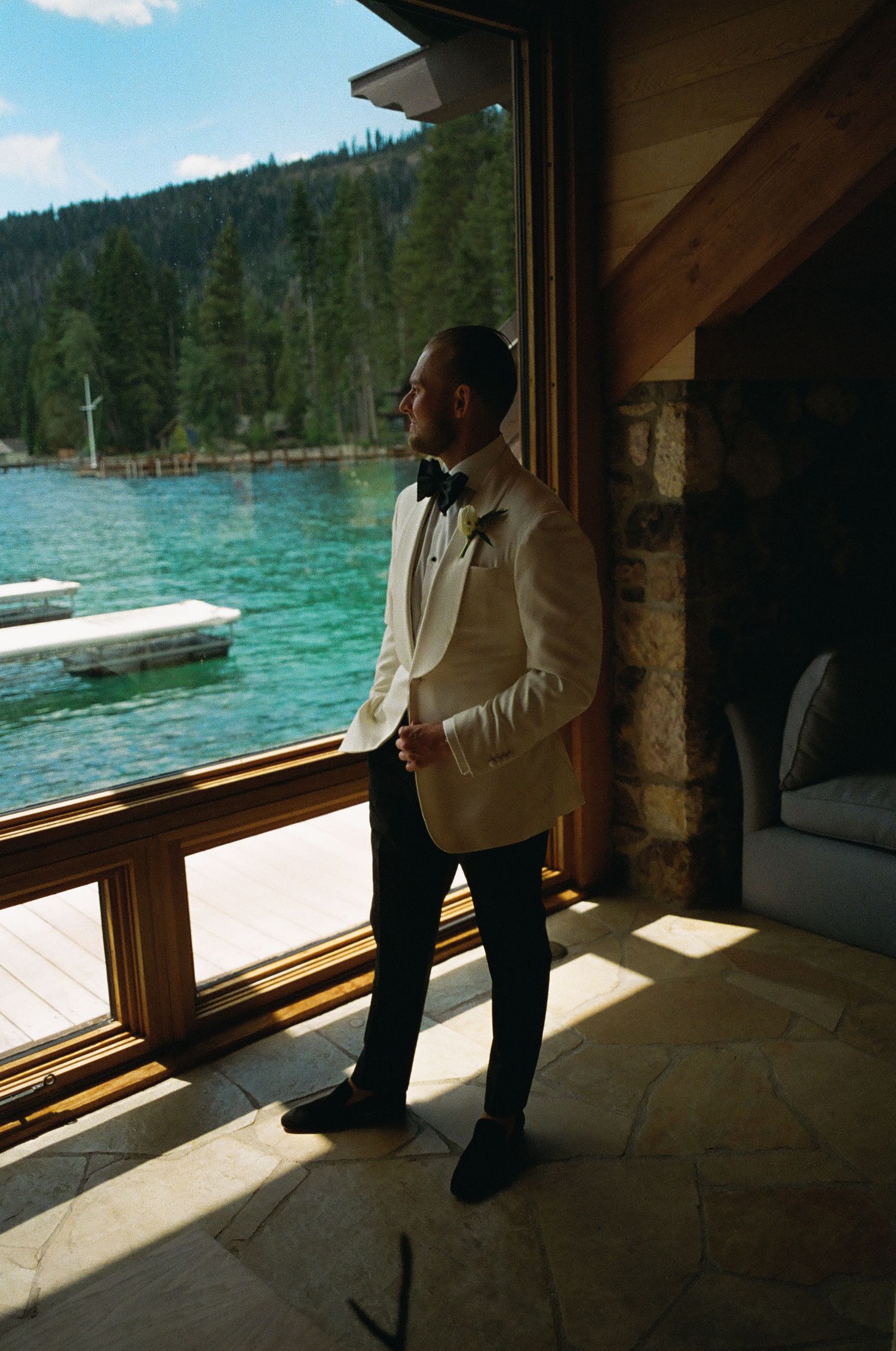 Silhouette of a person in formal attire standing by large windows overlooking a turquoise lake.