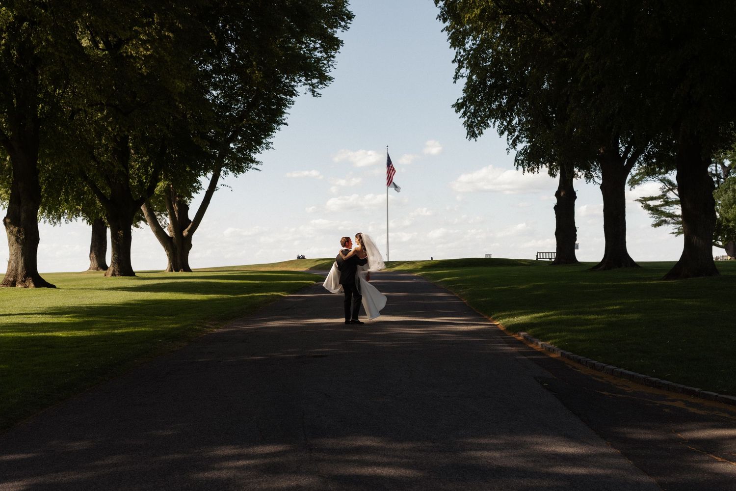 Couple embracing on a tree-lined path with an American flag in the background on a sunny wedding day.