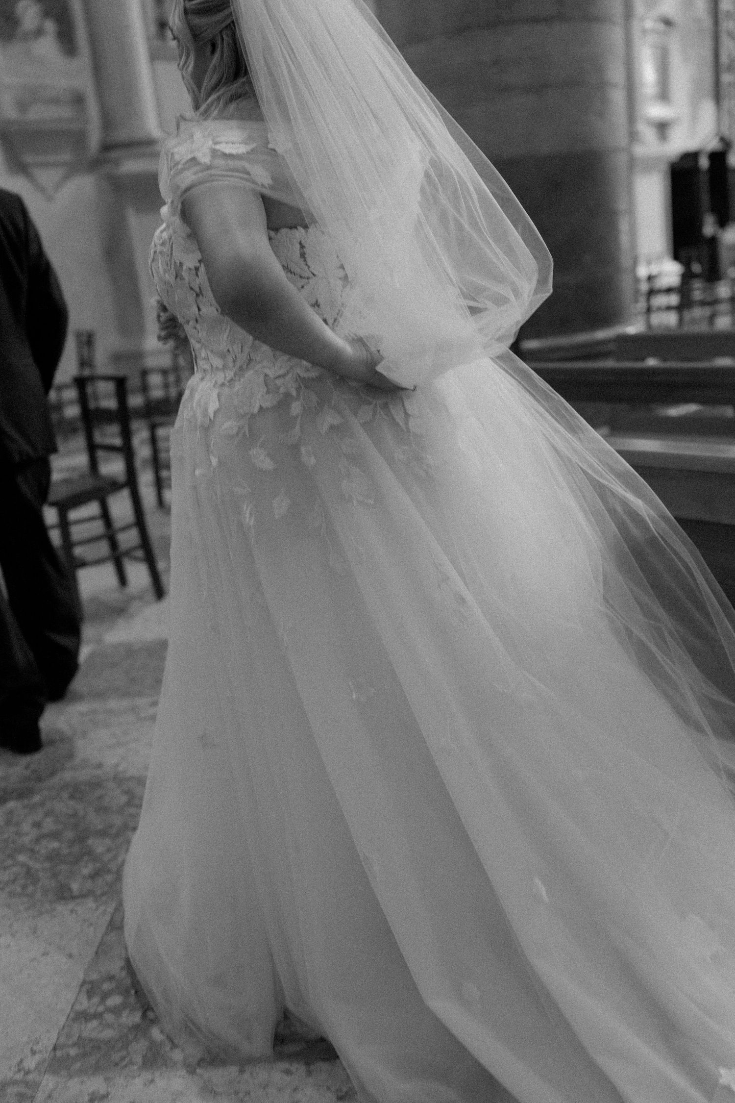 Black and white photo of bride in lace off-shoulder gown with flowing veil in church.