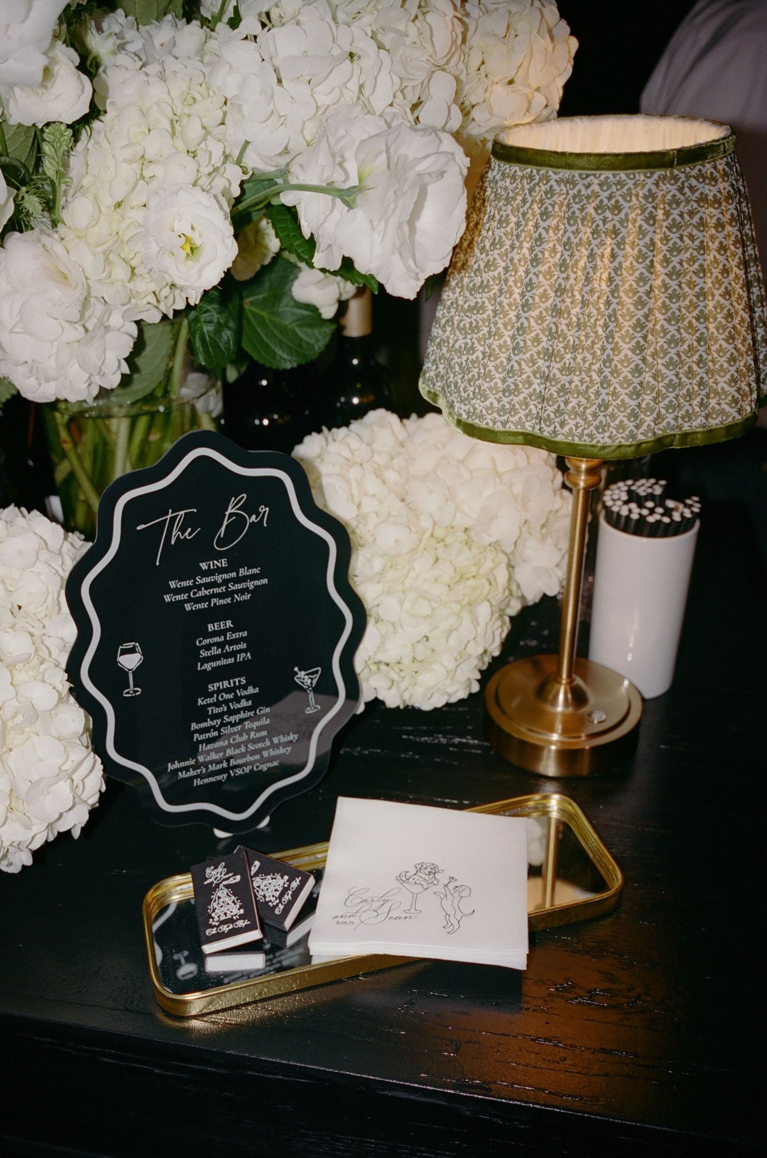 Wedding reception details including white flowers, menu sign, and gold table lamp on dark surface.