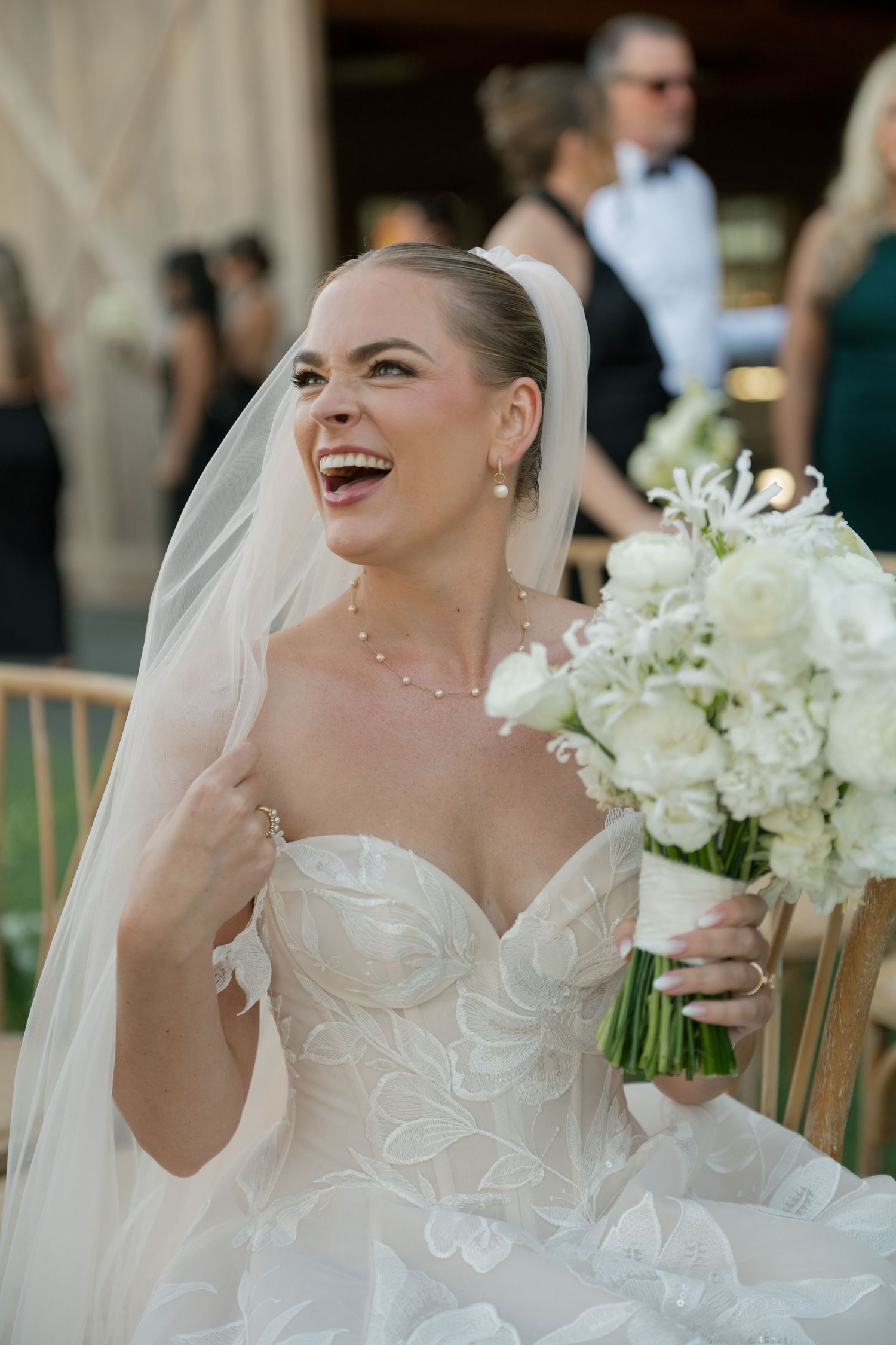 Bride in strapless lace gown holds white bouquet while laughing.