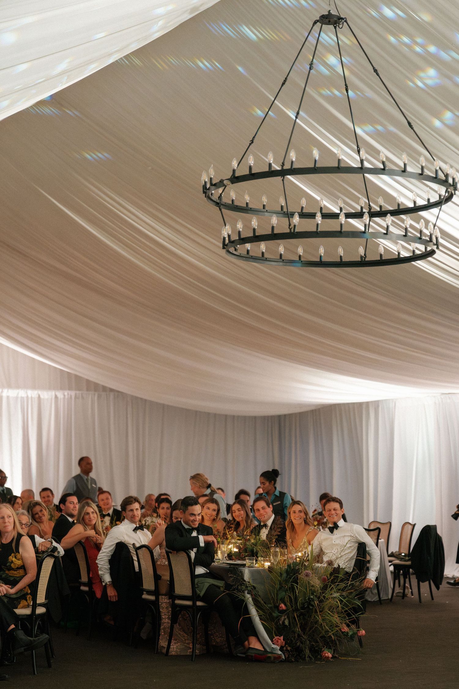 Wedding reception venue with circular chandeliers and white draping illuminated for evening celebration.