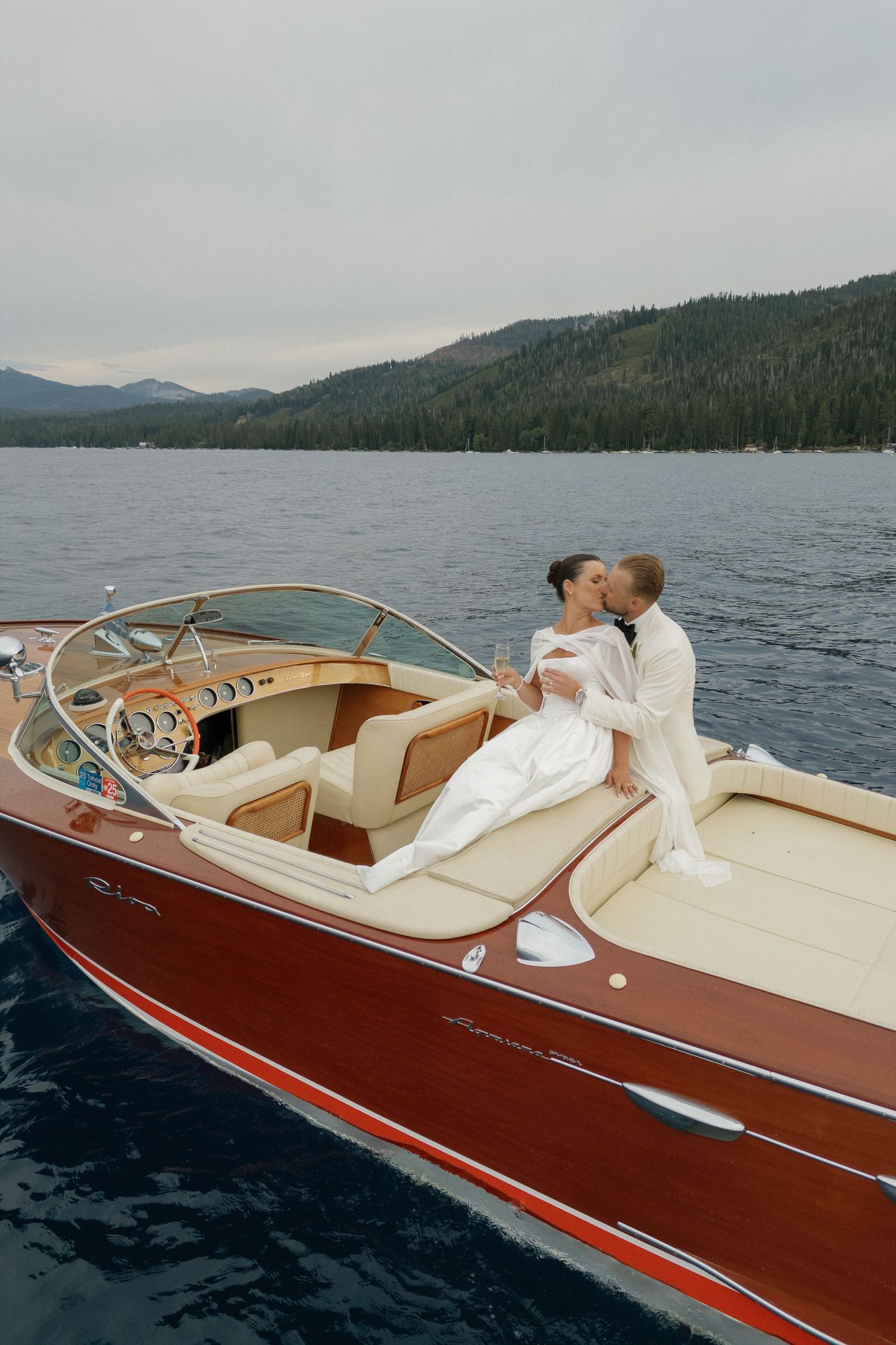 A classic wooden speedboat glides across a mountain lake on a cloudy day.
