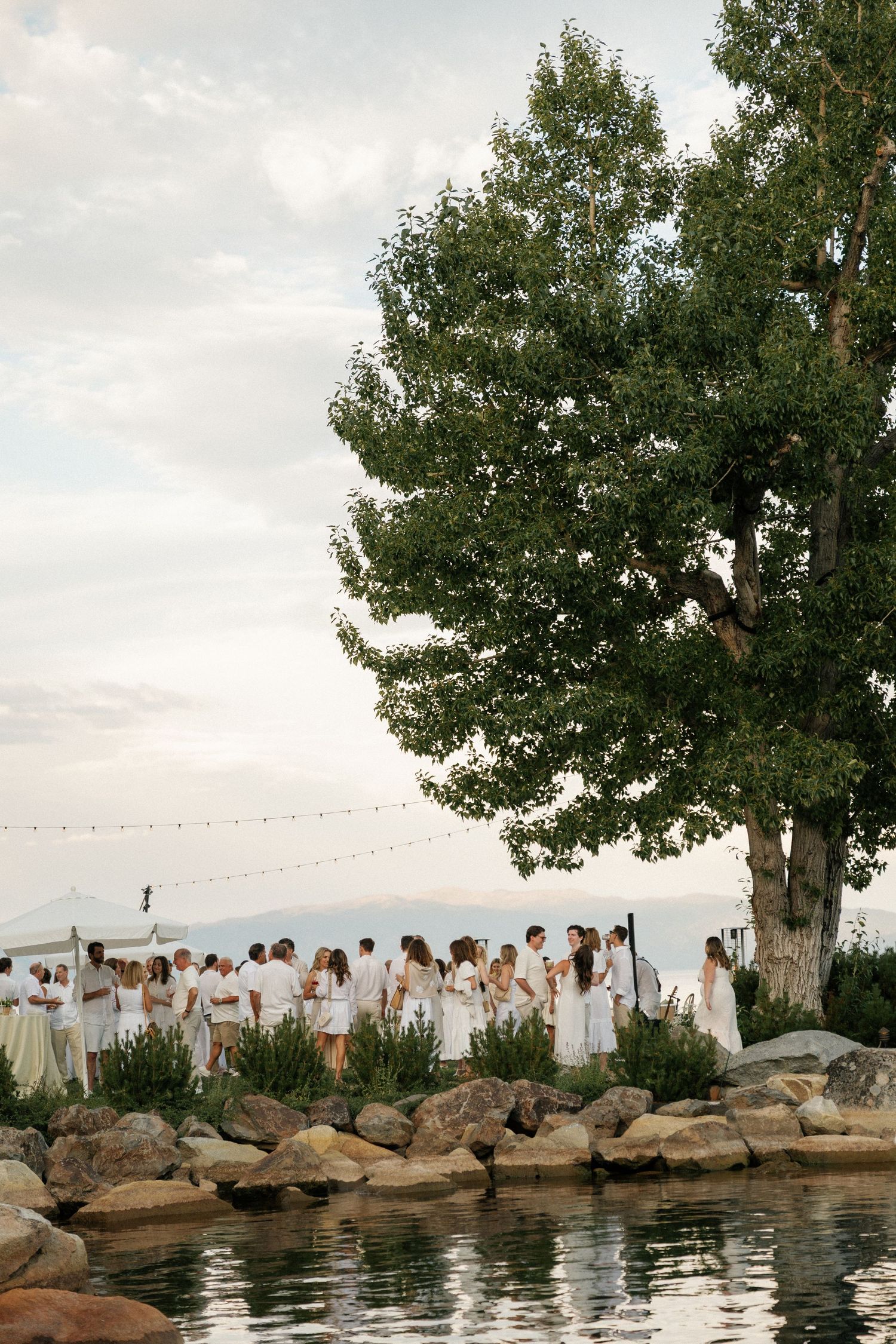 Wedding guests in white gather along rocky waterfront under string lights and trees.