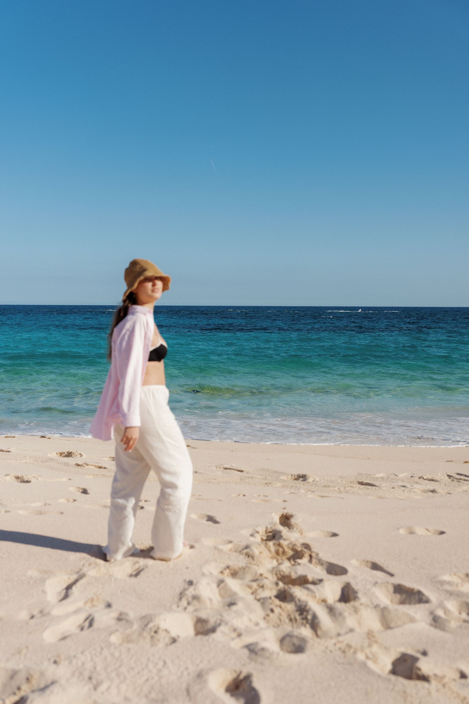A person in white clothing walks along a sandy beach with clear blue water in the background.