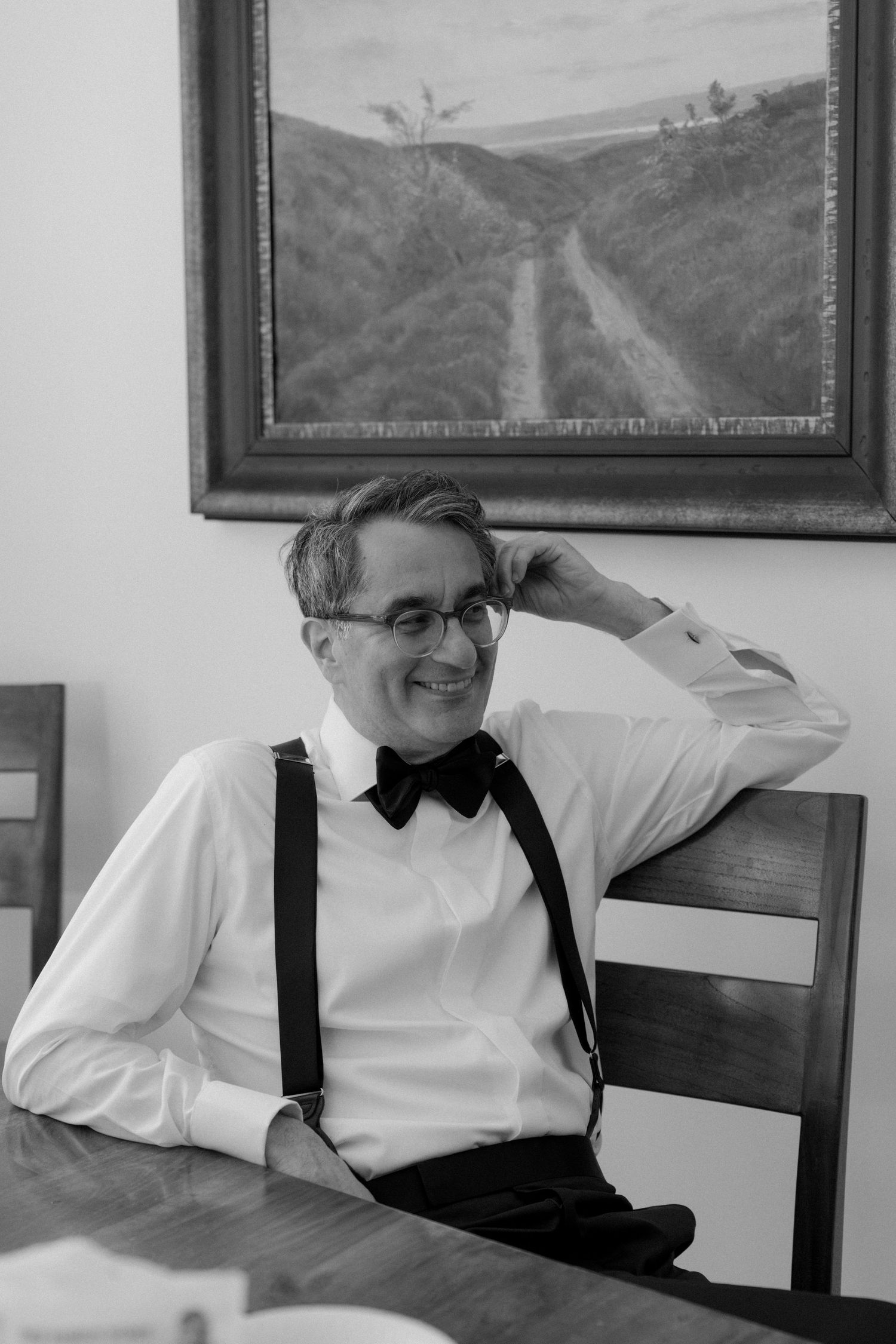 Man in white shirt, bow tie, and suspenders smiling while seated at a wooden table with a landscape painting behind him.