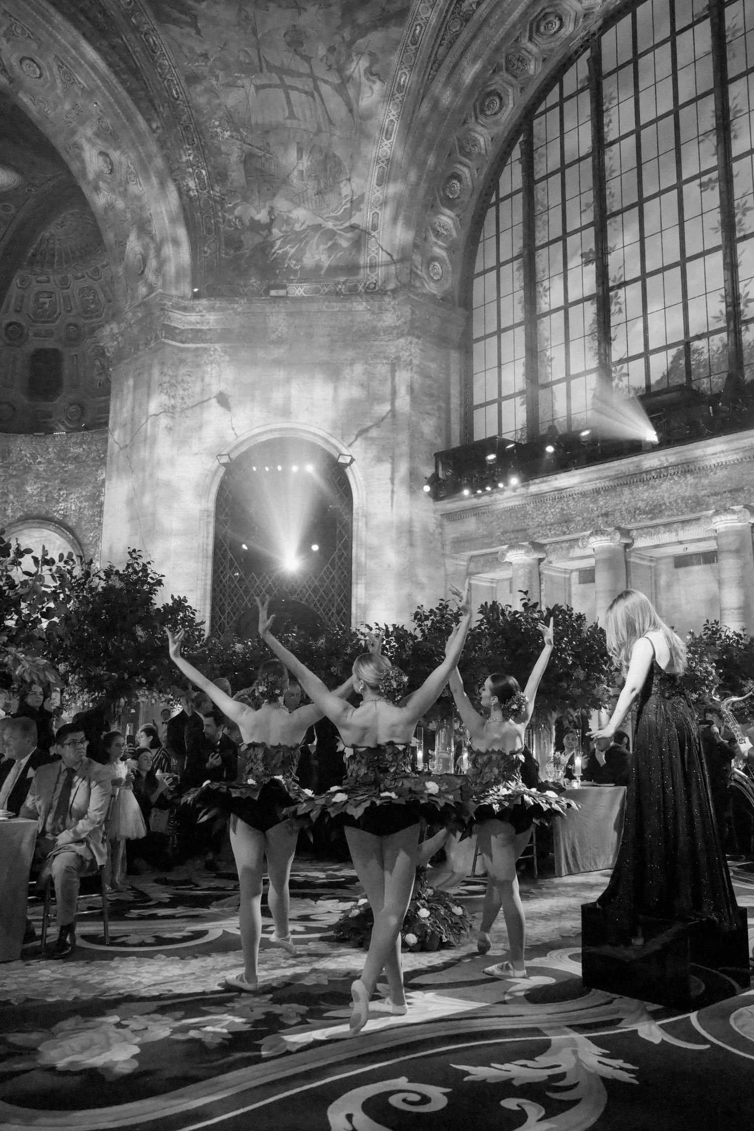 A black and white image of a grand, ornate interior with people gathered and performers on a stage.
