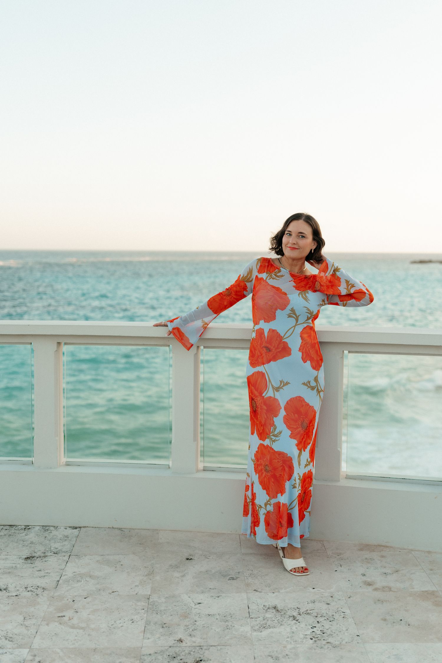 A woman in a bright red and white floral dress stands on a balcony overlooking the ocean.